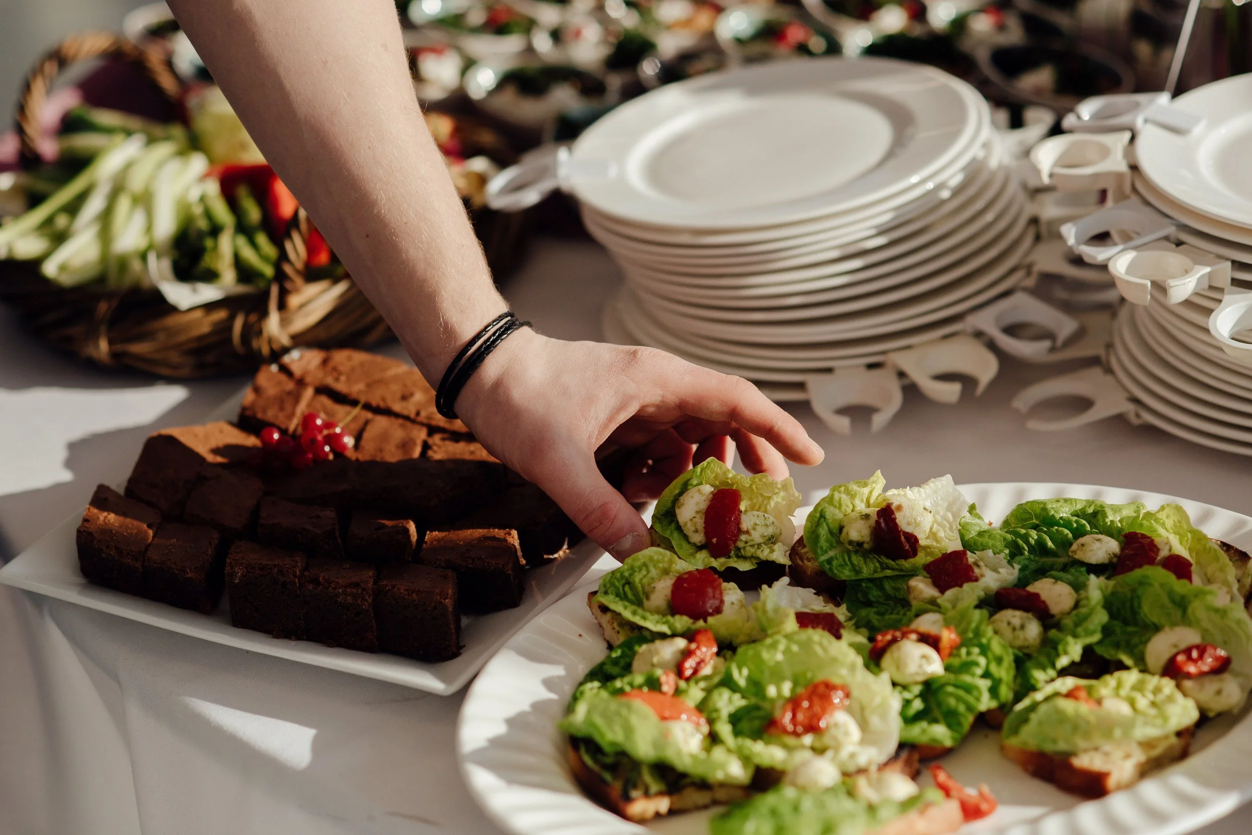 Person serving food at a buffet table with plates of salad, chocolate cake, and fresh vegetables.