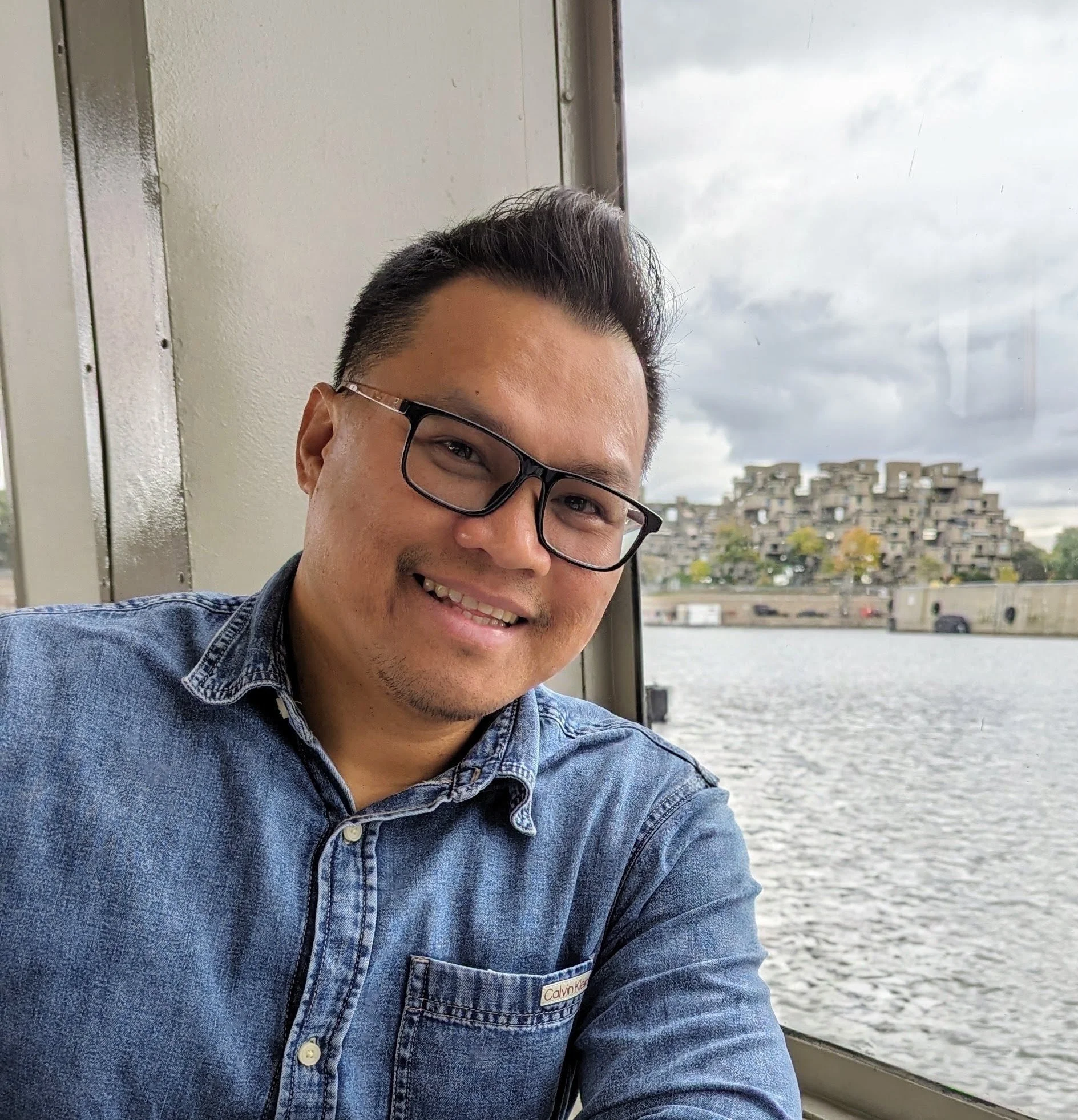 A man with glasses smiling for a selfie on a boat, with a river and buildings in the background on a cloudy day.