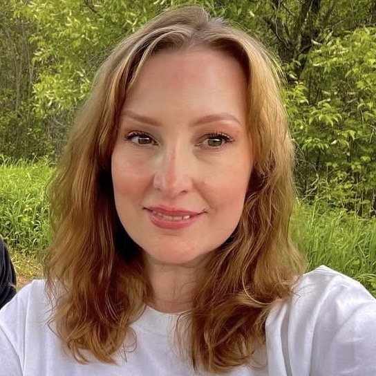 A woman with shoulder-length wavy red hair, wearing a white shirt, smiling outdoors with greenery in the background.