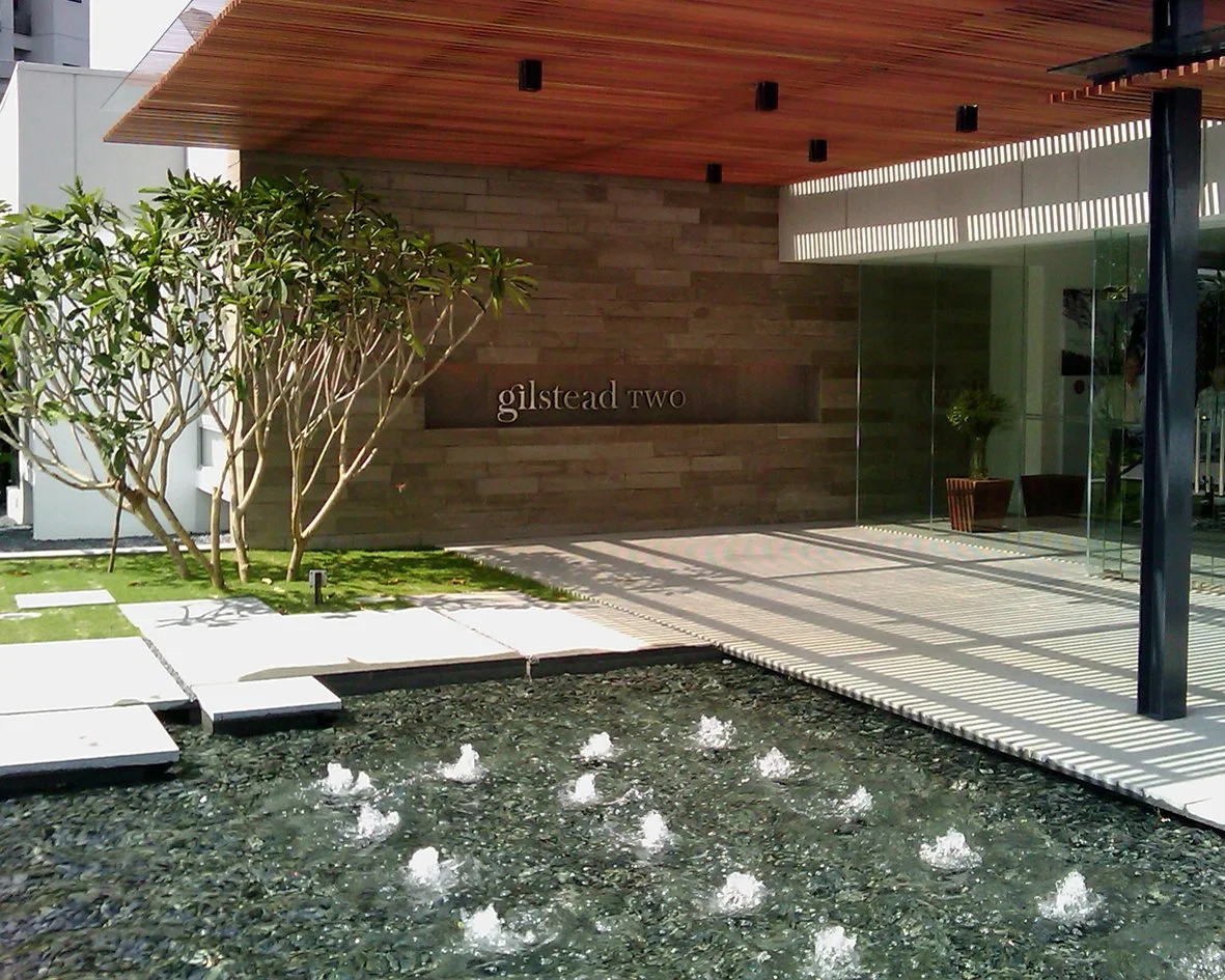 Modern building entrance with a water feature, a tree, and a wooden overhang. The building has a sign that reads 'gilstead two' and features glass walls, outdoor seating area, and decorative shadows.