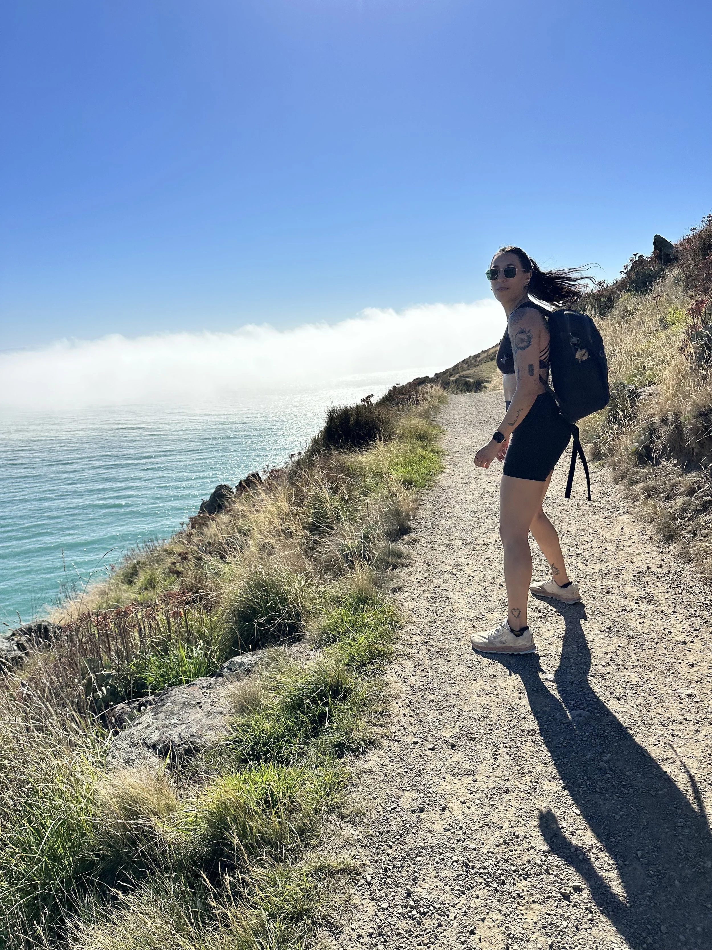 A woman with tattoos and sunglasses walking on a trail along a coastal cliffside with green vegetation, facing away from the camera, with the ocean and sky in the background.