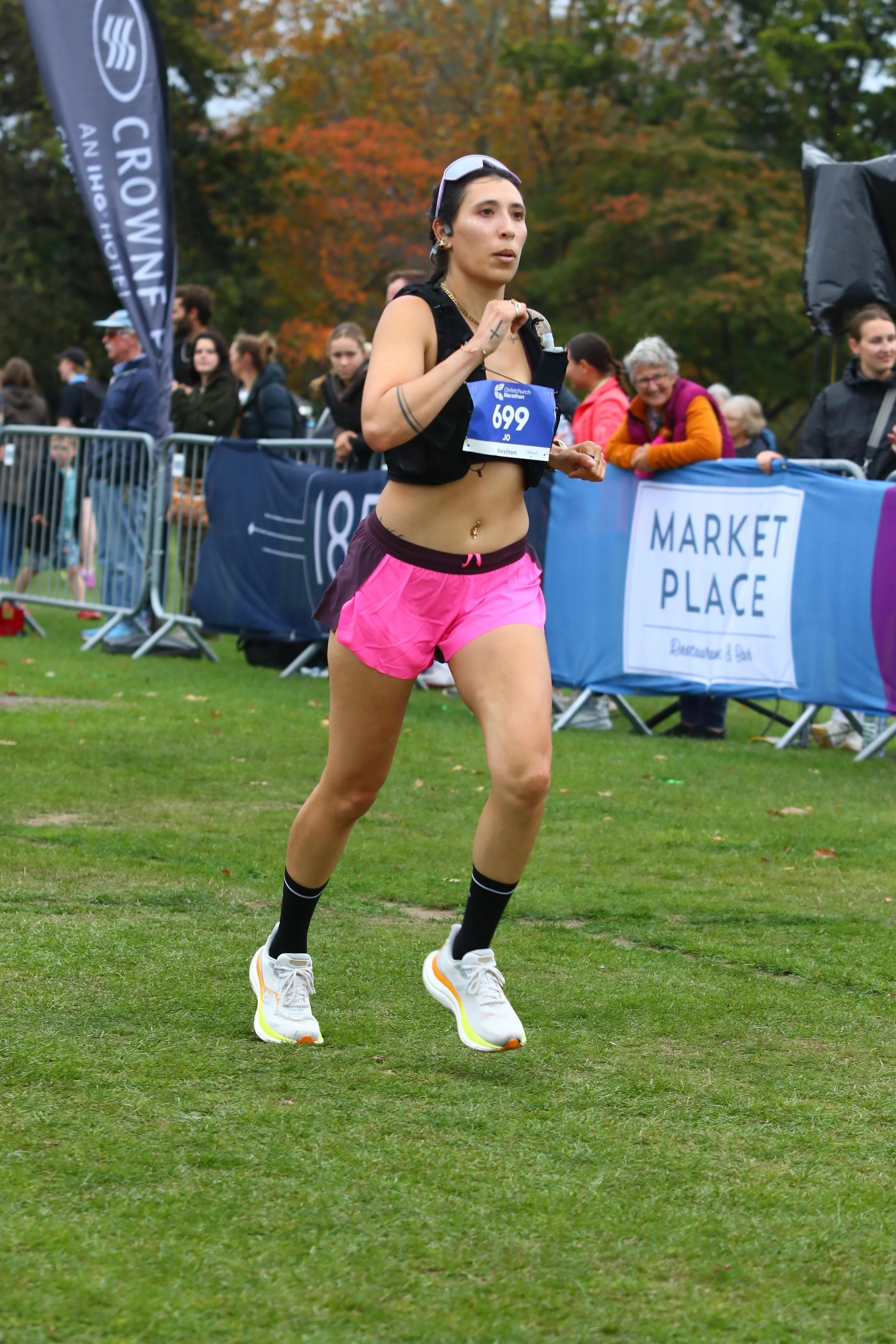 A female marathon runner in pink shorts and white running shoes runs on grass during a race, with spectators and race banners in the background.
