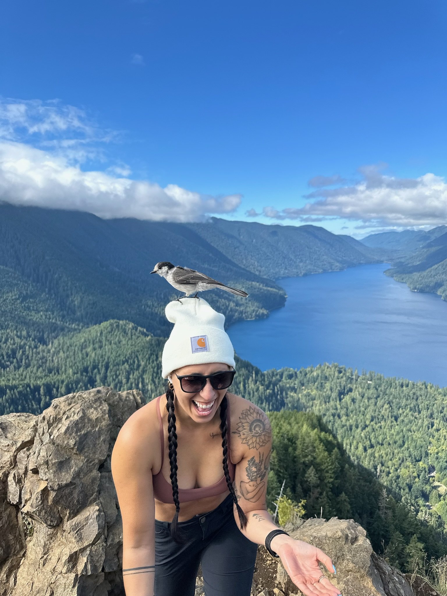 A woman wearing sunglasses, a white beanie, and a tank top smiling and laughing while sitting on a rocky ledge outdoors. A bird is perched on her head, and a scenic view of mountains, forests, and a river is in the background.