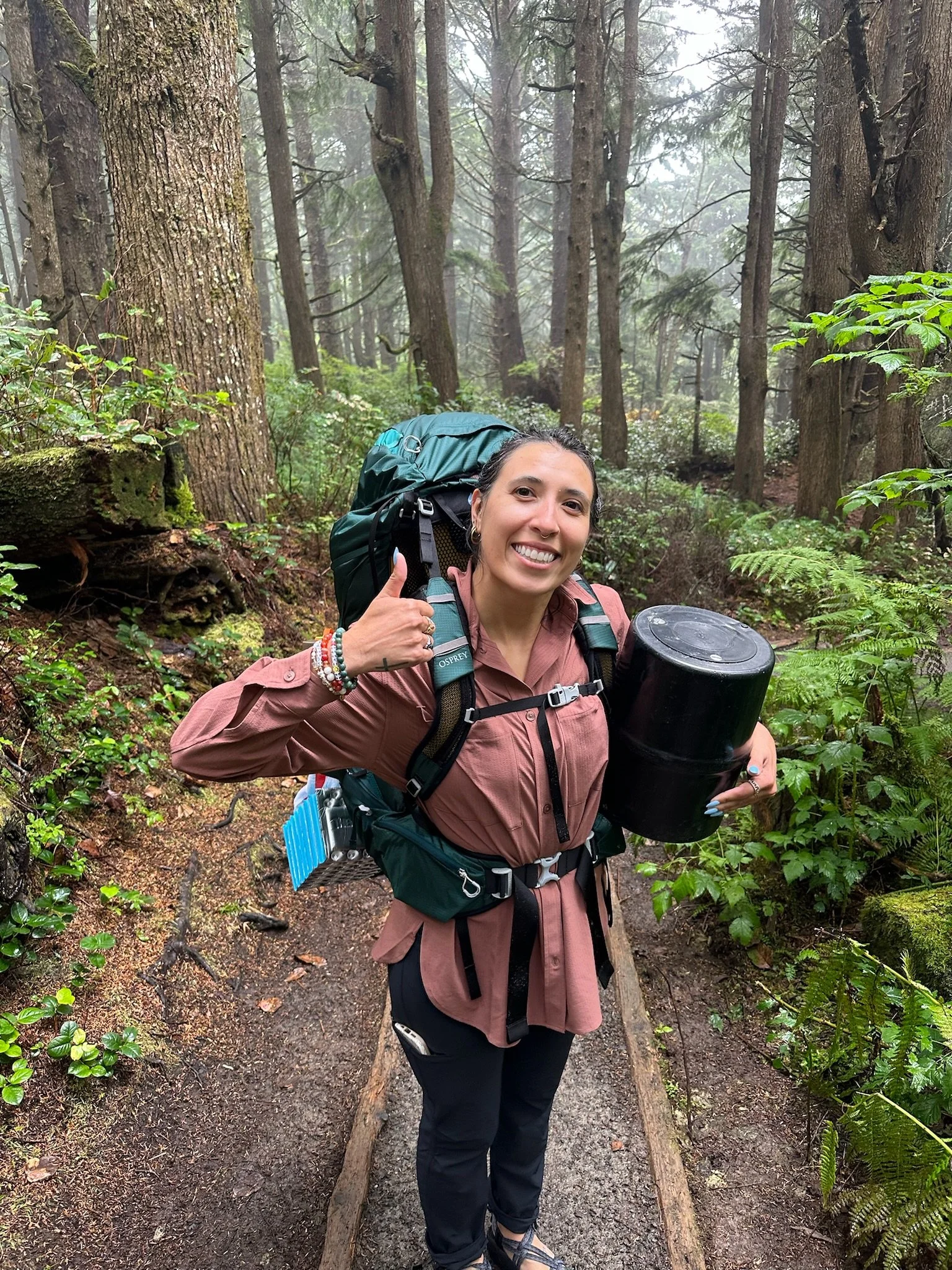 A woman smiling while hiking in a forest, giving a thumbs up, wearing a pink jacket, black pants, hiking backpack, and holding a black thermos.