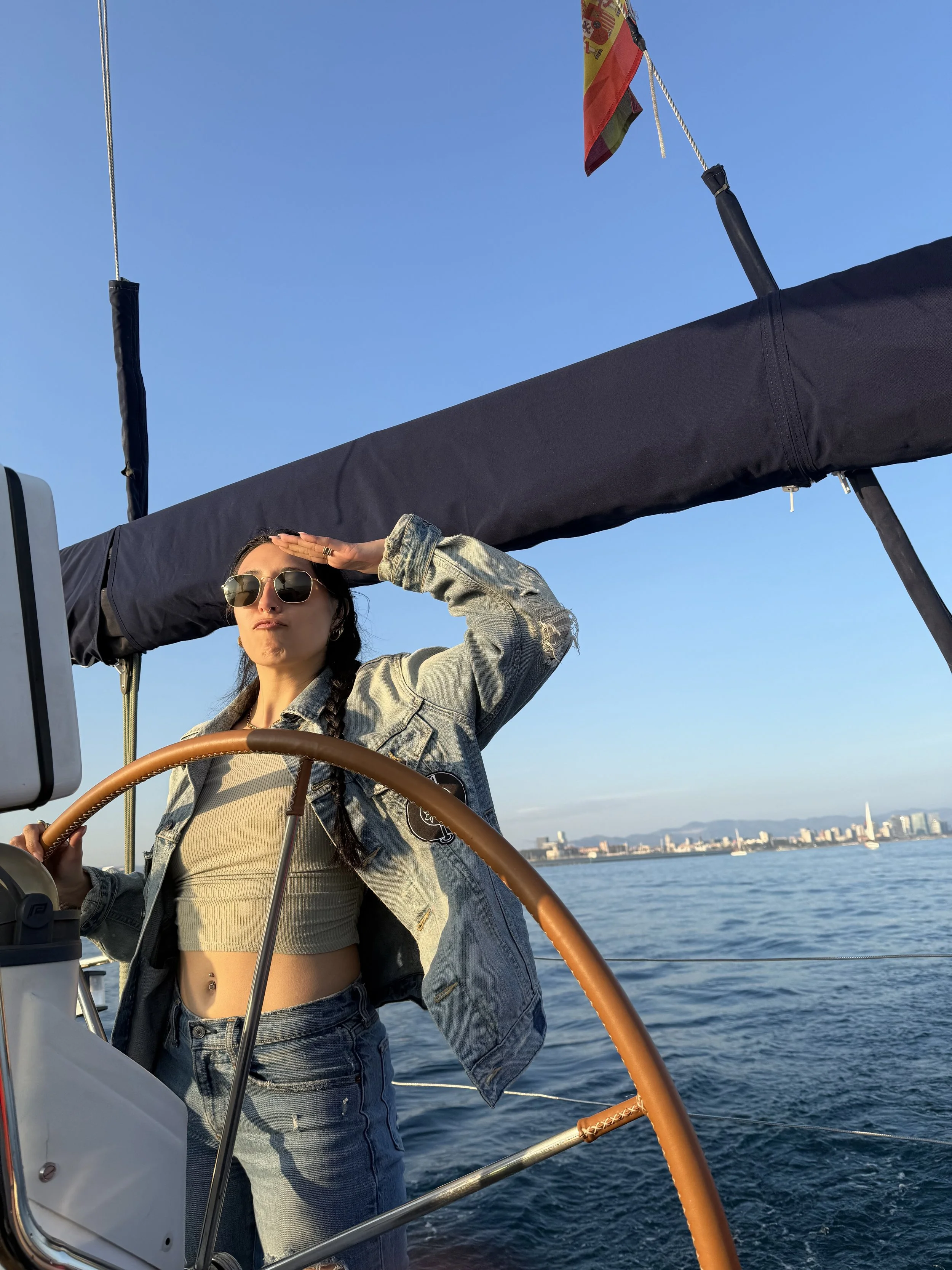 Woman on a sailboat, wearing sunglasses and a denim jacket, saluting with her hand, with the city skyline and water in the background under a clear blue sky.