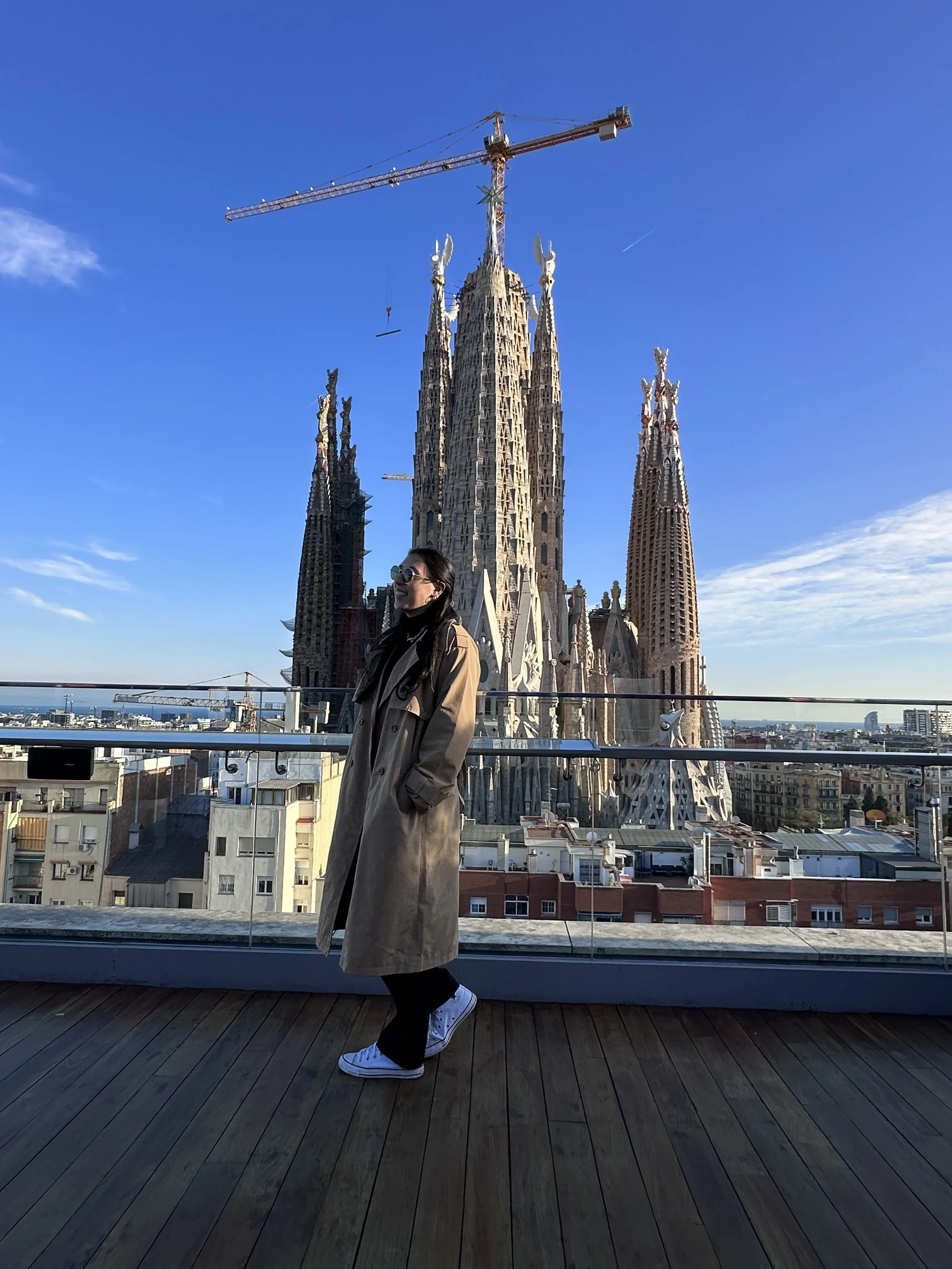 A woman standing on a rooftop terrace overlooking Barcelona with the Sagrada Familia cathedral in the background, wearing a trench coat, sunglasses, and sneakers.