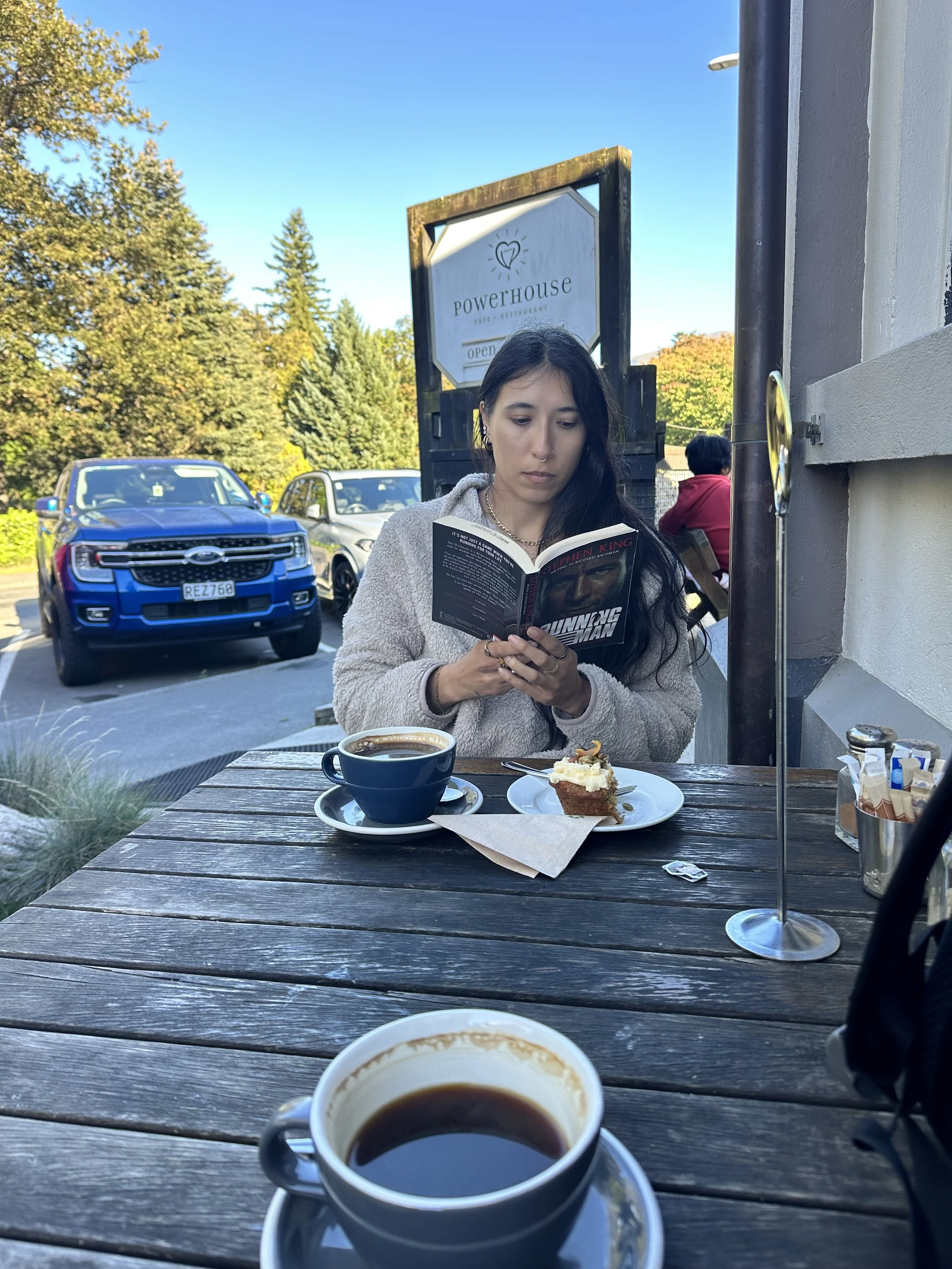 A woman with dark hair and wearing a beige jacket is sitting at an outdoor wooden table. She is reading a book and in front of her are a cup of black coffee and a slice of carrot cake on a plate. There is a signboard behind her that reads 'Powerhouse Cafe & Restaurant open,' and a parking lot with cars, including a blue Ford and silver vehicle, is visible in the background alongside trees with autumn foliage.