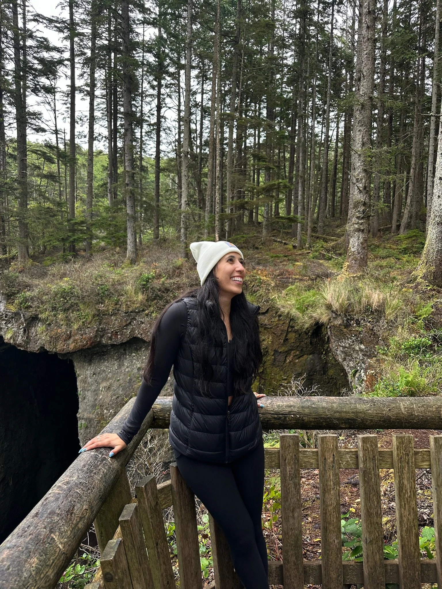 A woman smiling and wearing a white beanie and black puffer jacket standing outdoors in a wooded area with tall trees and a wooden fence.