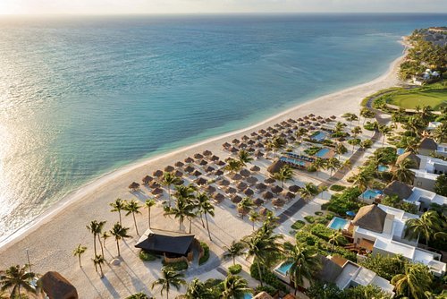 Aerial view of a beach resort with sun umbrellas, lounge chairs, palm trees, and ocean.
