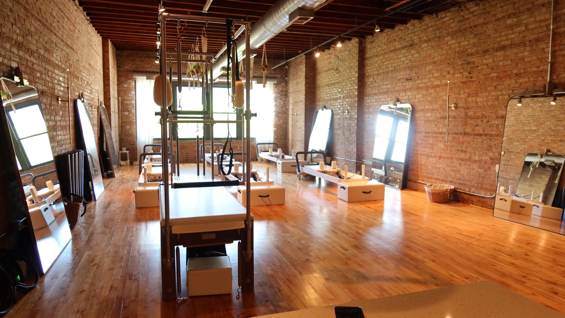 Interior of a Pilates or yoga studio with brick walls, wooden flooring, large windows, and various exercise equipment including mirrors, mats, and workout apparatus.