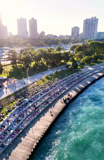 A city park with trees, a river with a curved walkway along its edge, and a shoreline with parked cars and outdoor seating. In the background, tall city buildings are visible under a bright sky.
