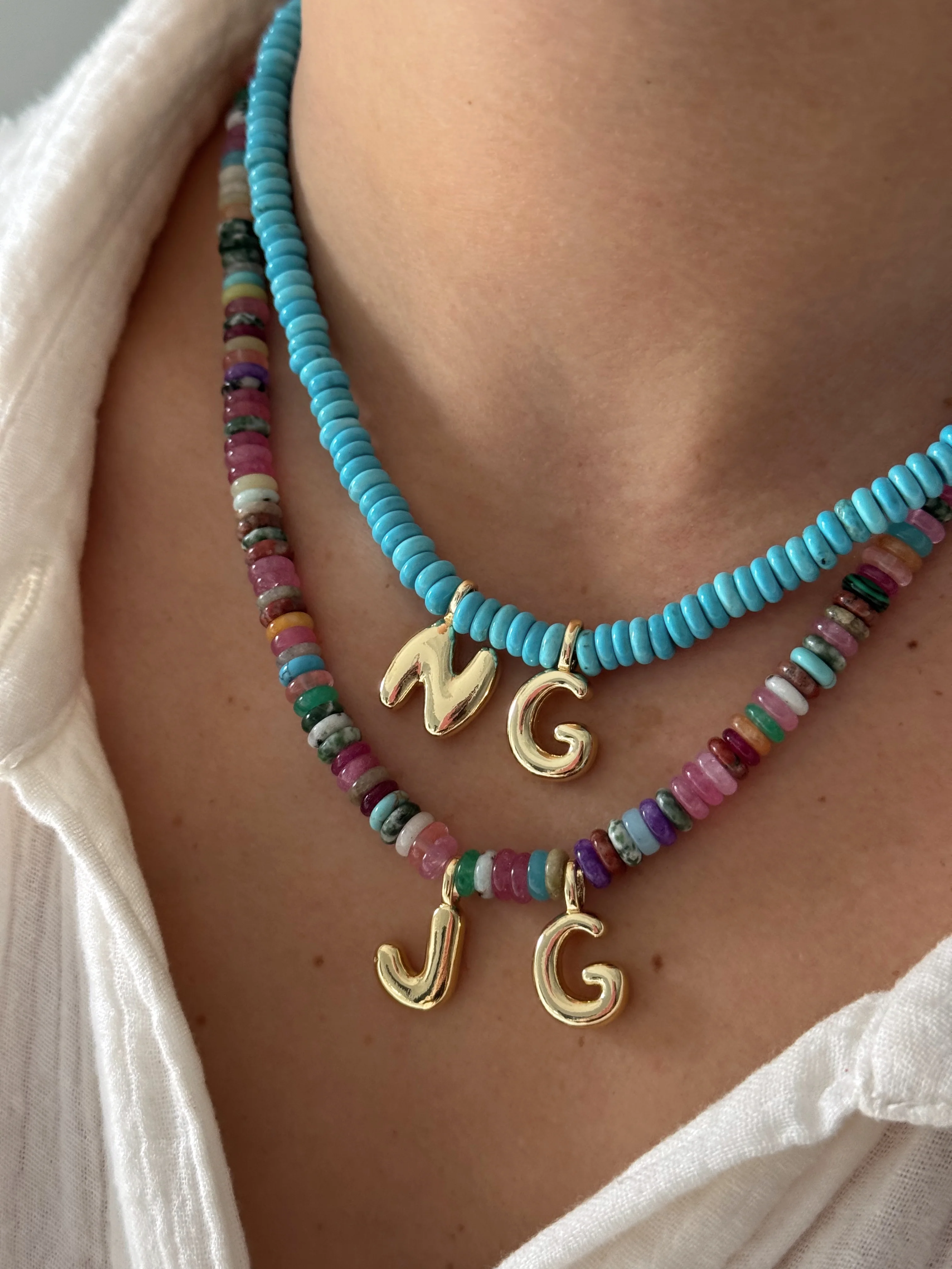 Close-up of layered beaded necklaces with gold initial charms on a person wearing a white garment.