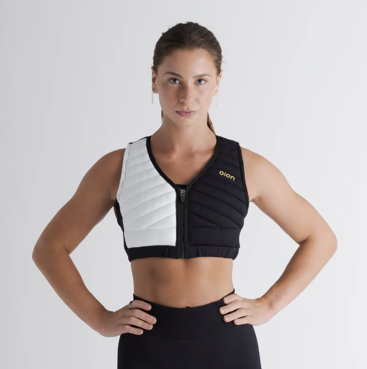 Woman wearing a black and white sleeveless sports vest with a zipper, standing with her hands on her hips against a plain white background.