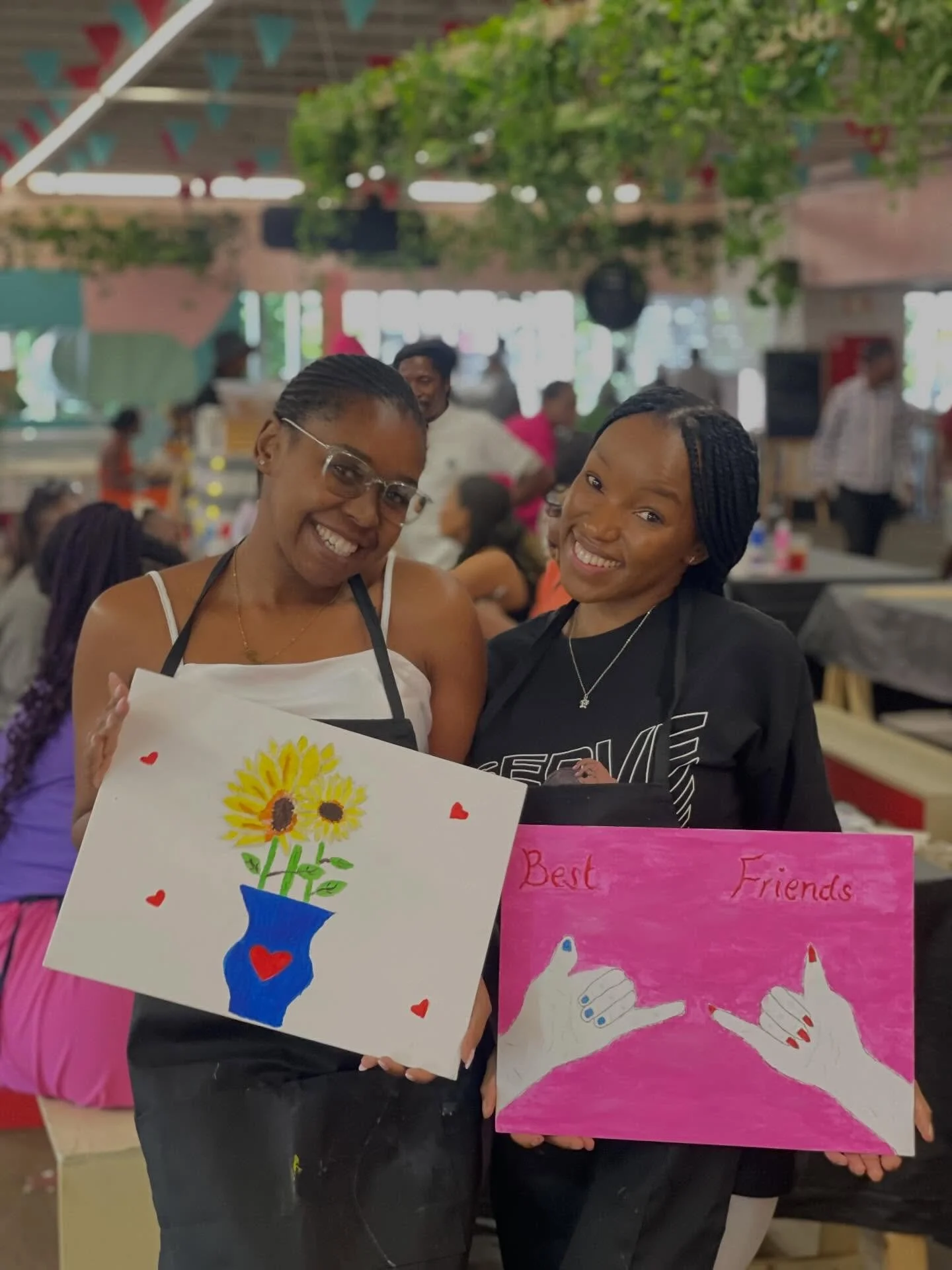 Two women smiling and holding handmade signs at an indoor event, with decorations and other people in the background.