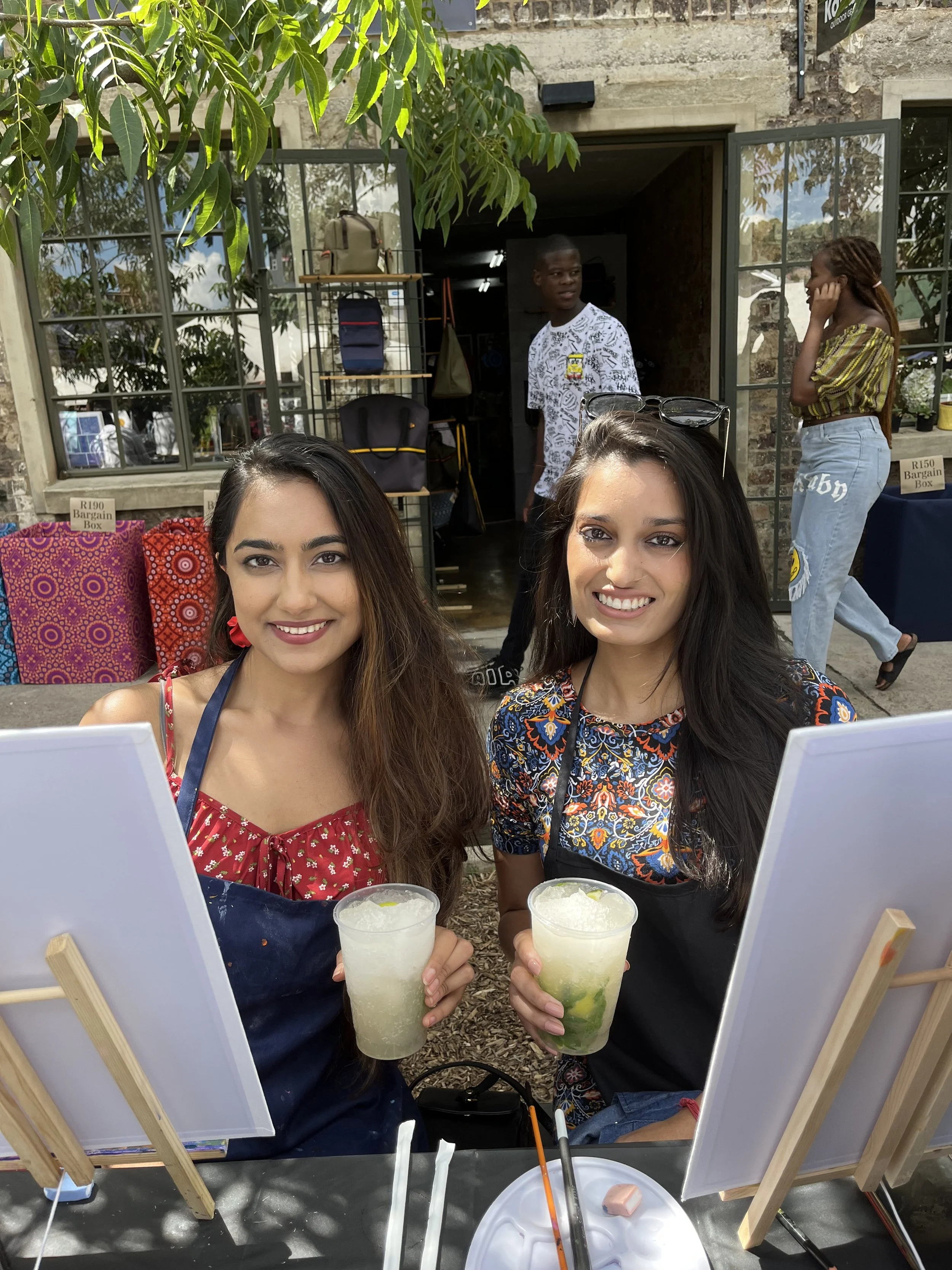 Two women smiling and holding drinks at an outdoor painting event, with easels and paint supplies in front of them, while a man and a woman are seen in the background near a building with jars of craft supplies and shelves.