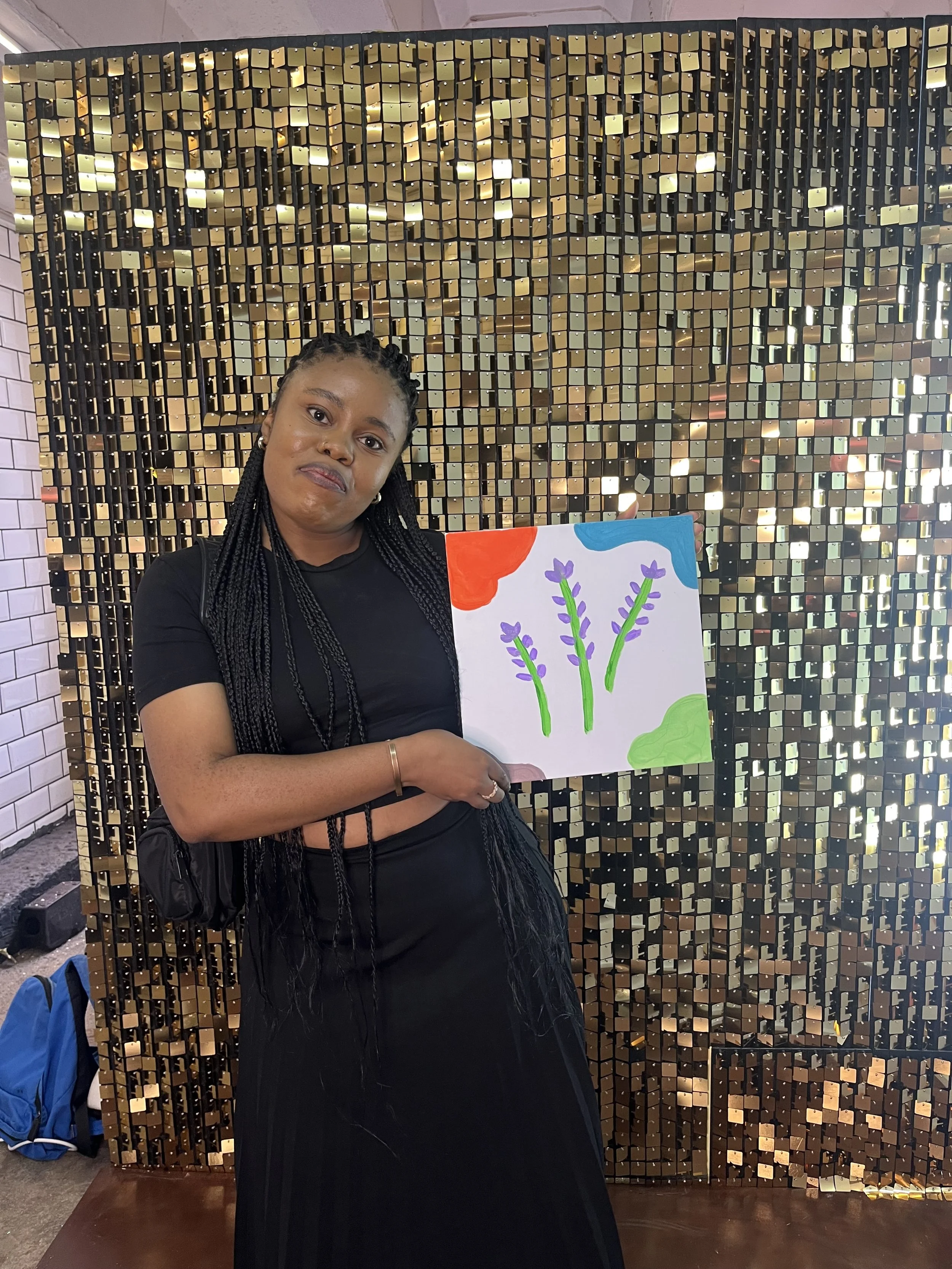 A woman with braided hair wearing a black shirt is holding a colorful drawing of flowers in front of a decorative wall with gold and black tiles.