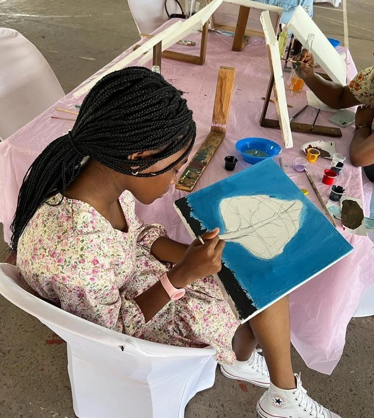 A young girl with braided hair is sitting on a white chair, painting on a canvas. She is wearing a floral dress and a pink wristwatch. The canvas has a partly finished blue background with a white shape in the center. She is in an indoor or outdoor a