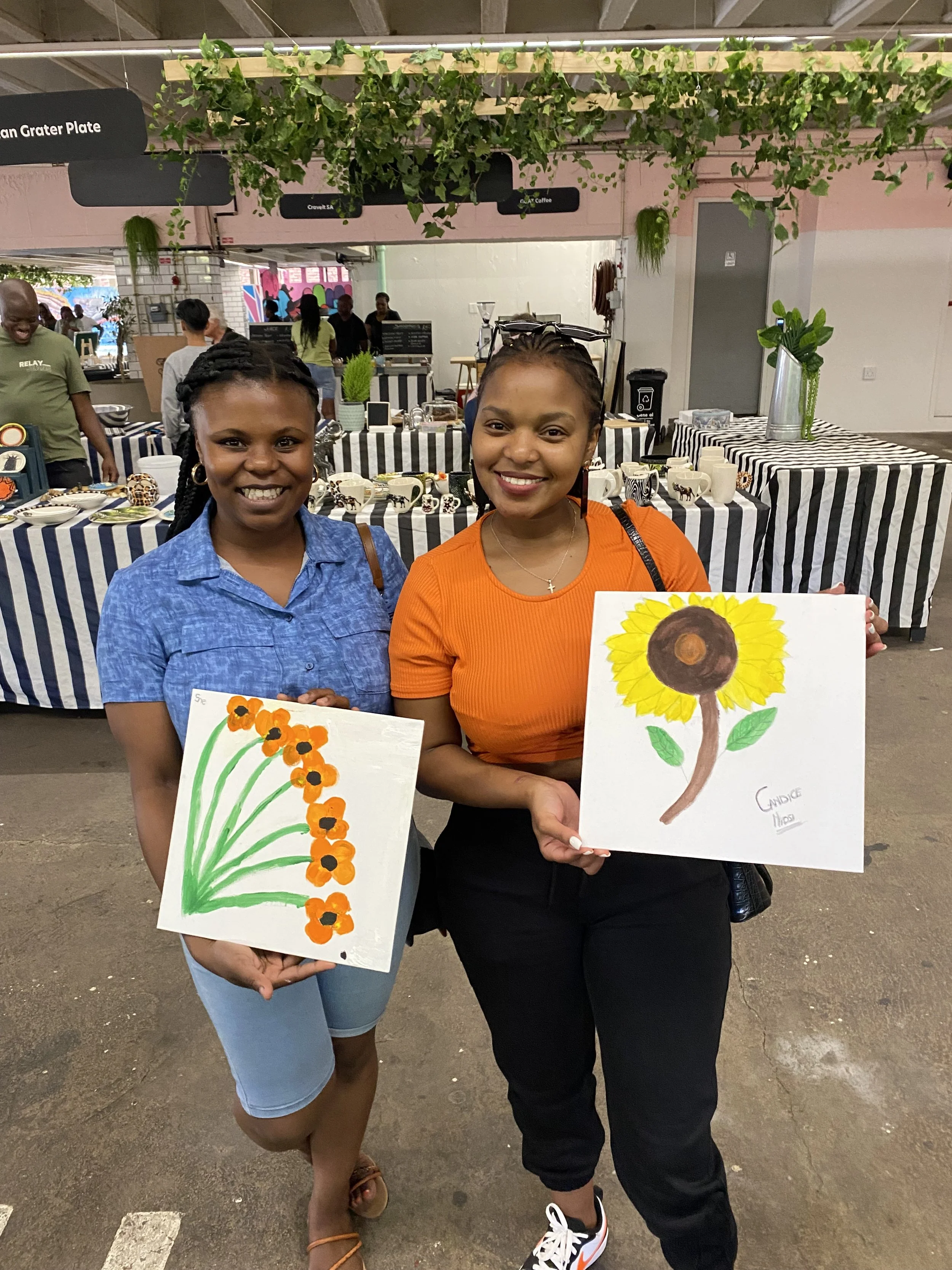 Two smiling women with dark hair and brown skin standing inside a building, each holding a colorful painting of flowers. The woman on the left is wearing a blue shirt and a denim skirt, while the woman on the right is wearing an orange top and black 