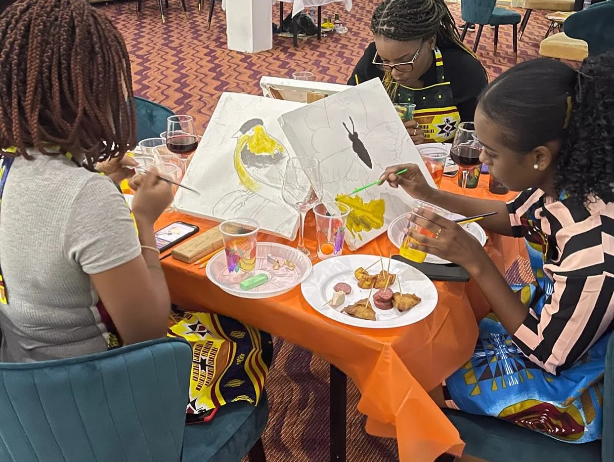 Three women painting a large canvas at a table during a social event. The table has snacks, drinks, and painting supplies, and they are focusing on creating artwork involving a parrot and a butterfly.
