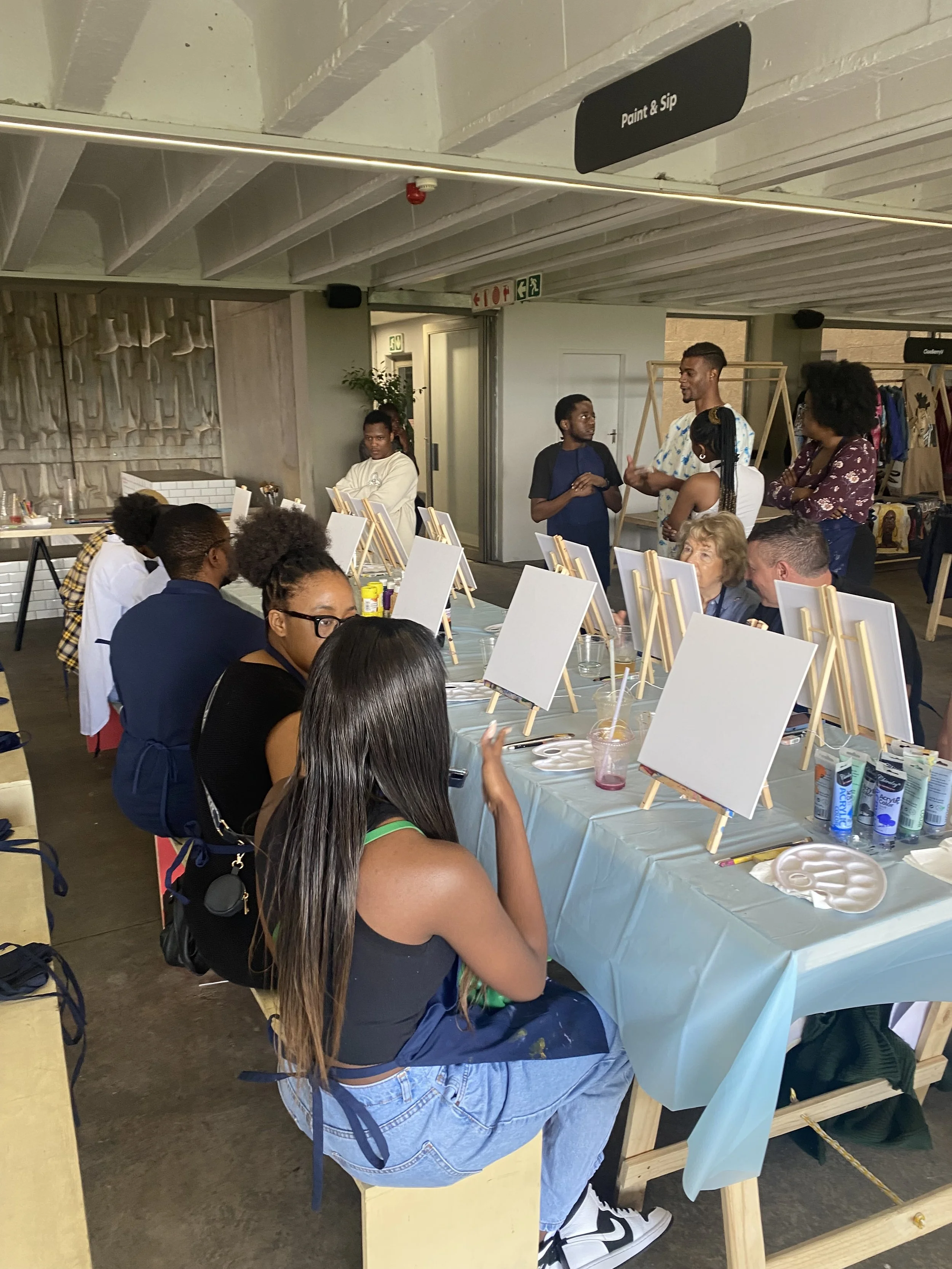People engaging in a painting activity at a table with canvases and art supplies in an indoor space, some people are sitting while others are standing and talking.