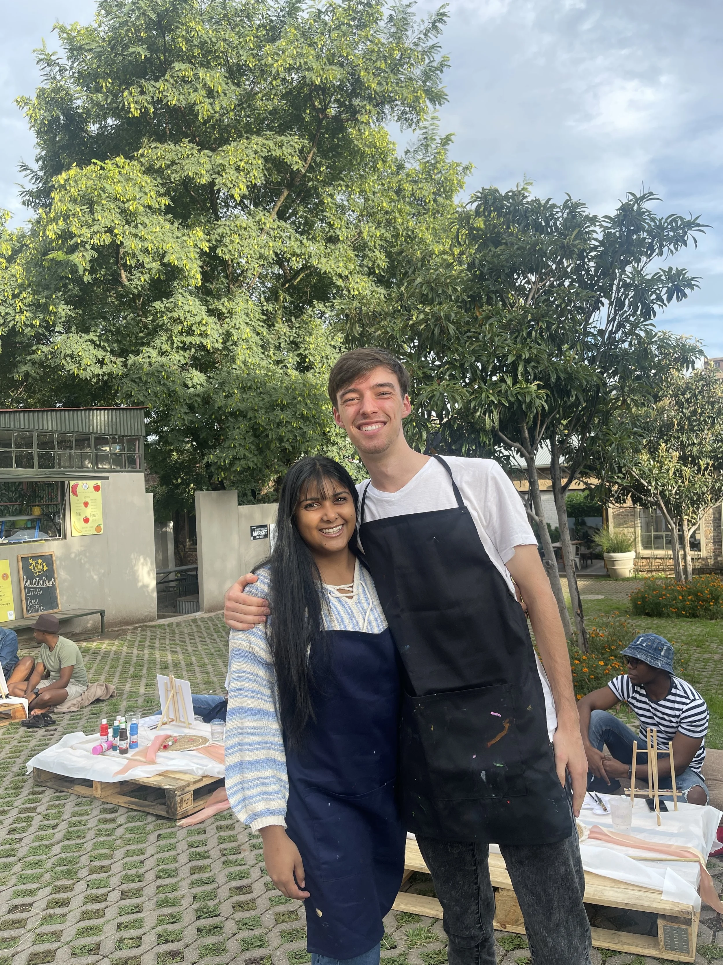 Two people smiling and posing for a photo outdoors, with trees and people sitting at tables in the background.
