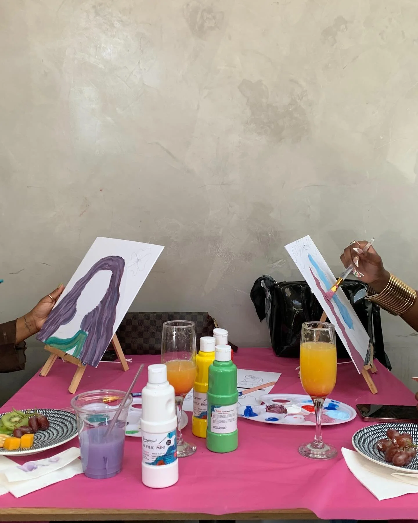 Two people painting colorful pictures on small easels at a table covered with a pink tablecloth. The table has several bottles of paint, drinks, and snacks, with a gray textured wall in the background.