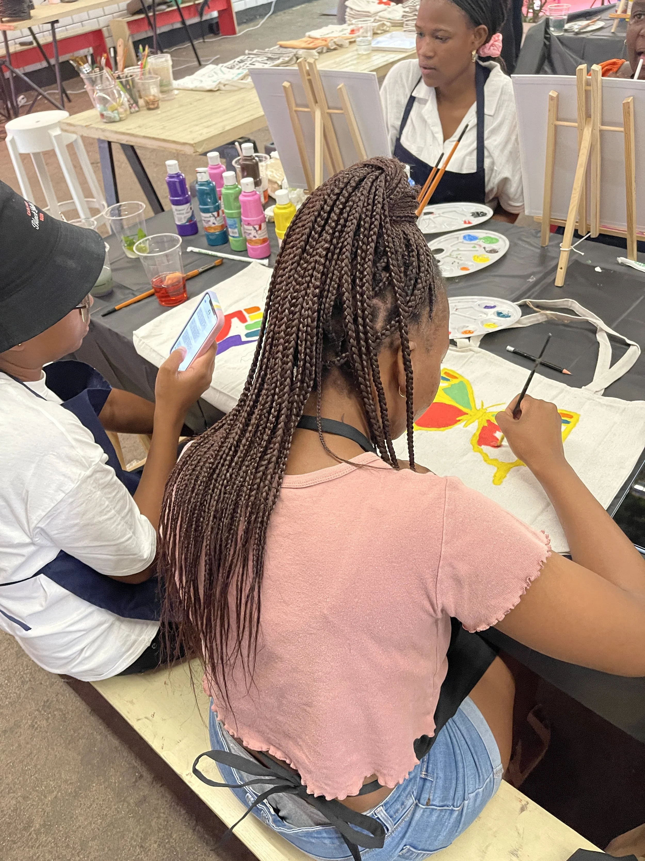 Three women are sitting at a table, painting colorful butterfly designs on fabric or paper with paint and brushes. One woman is absorbed in her work, another is looking at her phone, and the third is partially visible. The table has paint bottles, pa