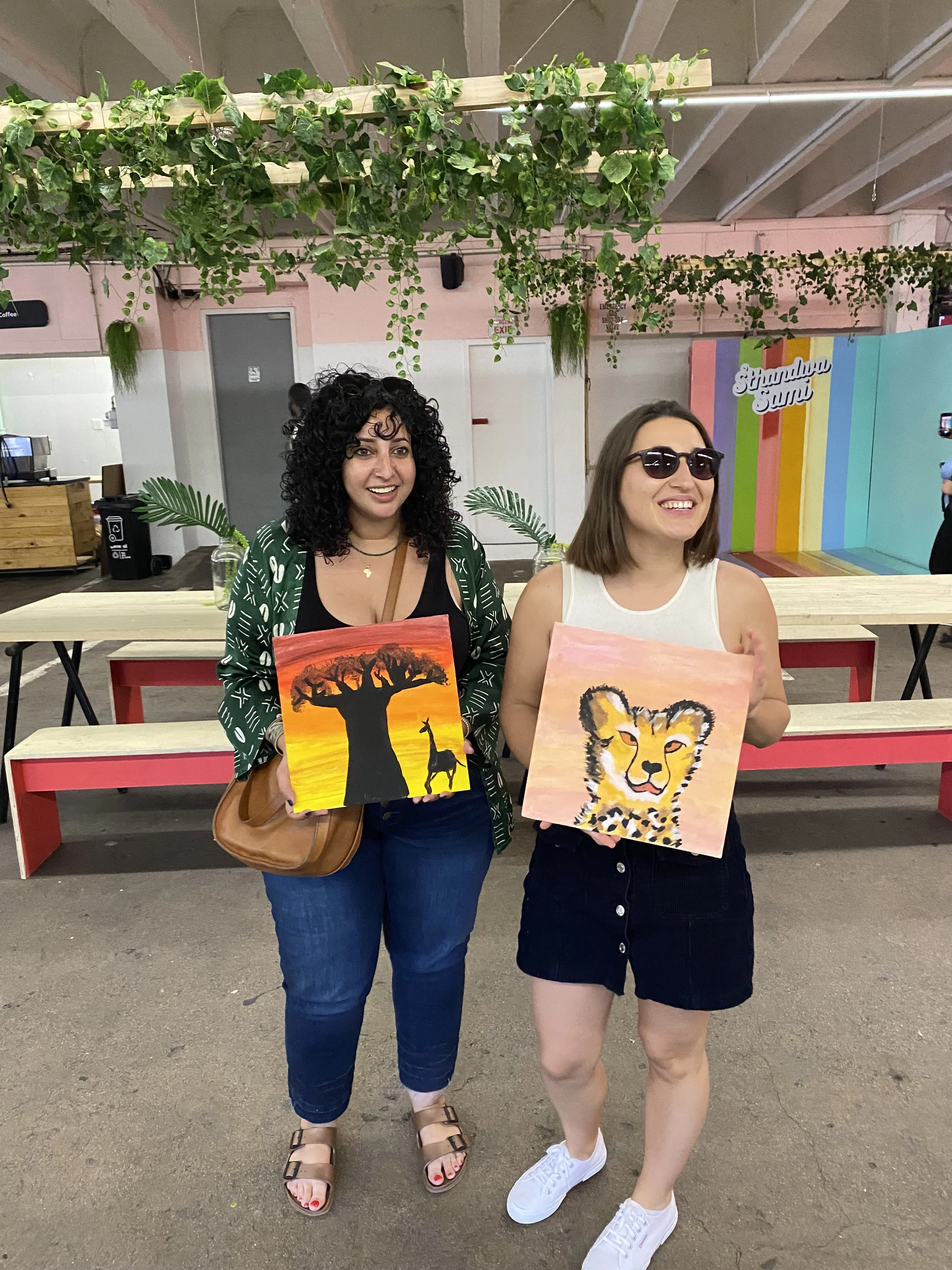 Two women standing side-by-side, each holding a painting. The woman on the left has dark curly hair, wearing a green patterned cardigan, dark jeans, sandals, and a necklace. The woman on the right has straight brown hair, wearing sunglasses, a white 