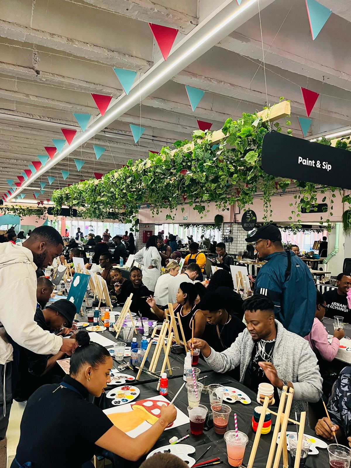 People participating in a group paint and sip event inside a decorated cafe with banners and greenery hanging from ceiling.