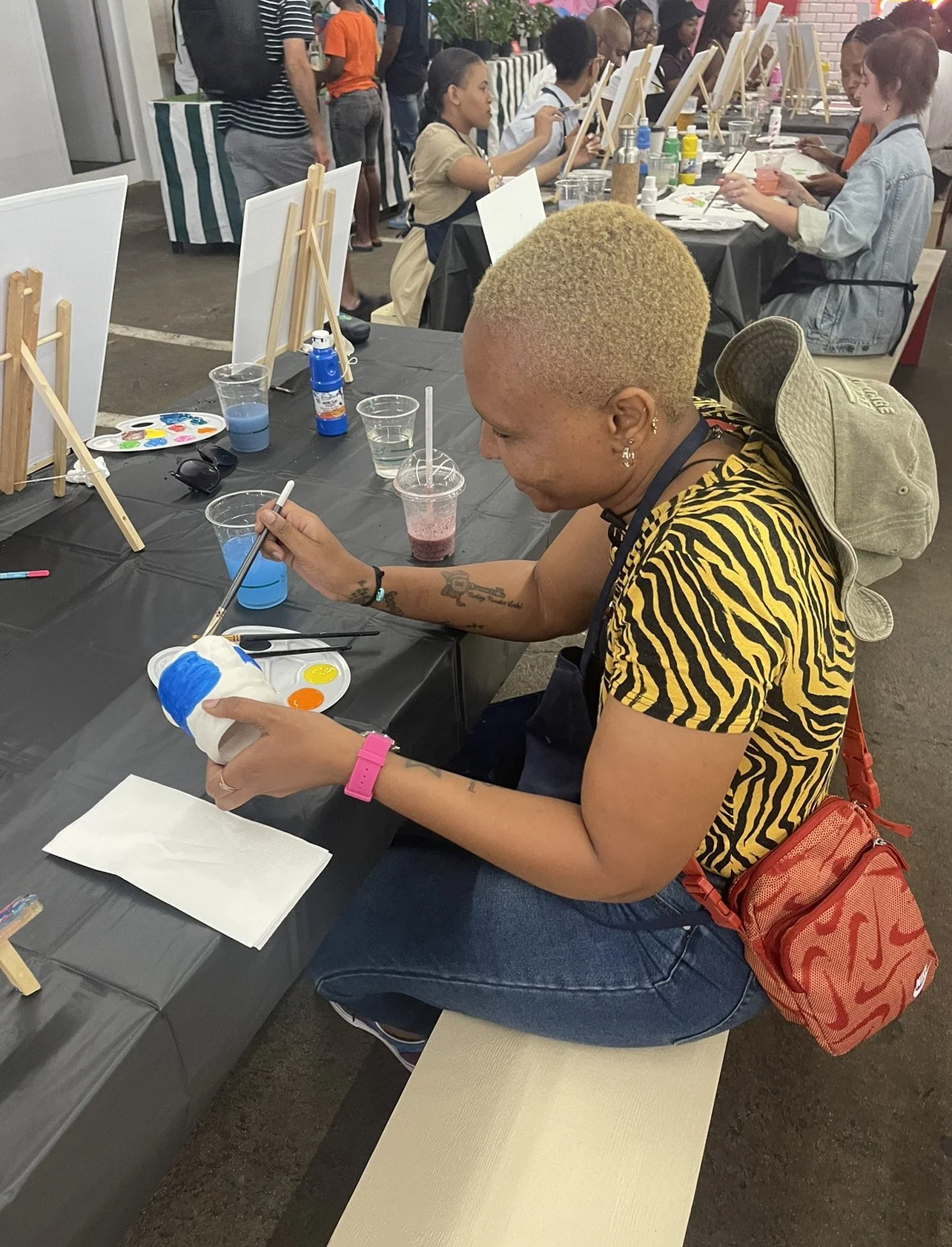 A person with short blond hair with a tiger stripe shirt painting a small sculpture. The person has tattoos, earrings, and a pink wristwatch. They are sitting at a table with paint supplies, and a backpack is hanging on their chair.