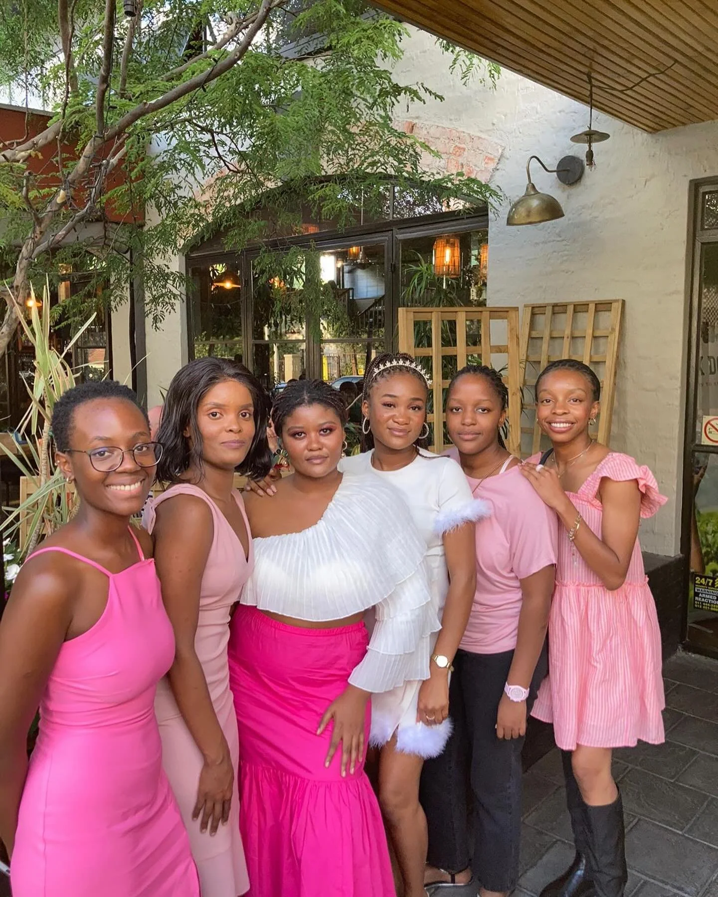Group of six women standing together outside, dressed in pink and white outfits, smiling for the camera.