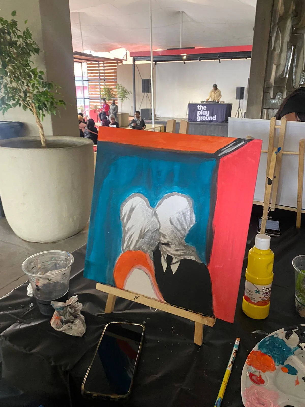 An art painting on an easel depicting two figures with angel wings facing each other, painted with a blue background and red on the sides, at an indoor art event with people and a DJ in the background.