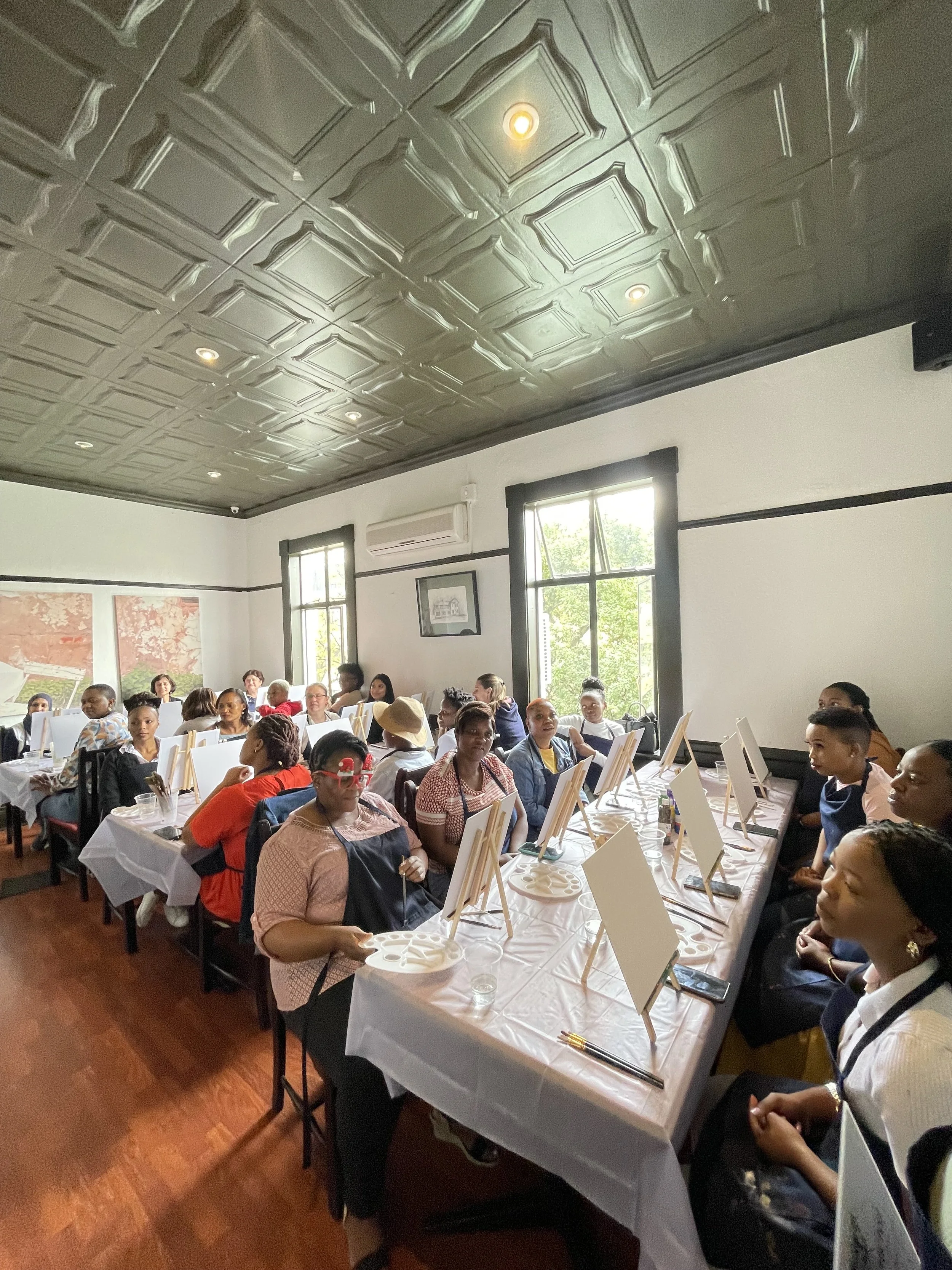 A group of people sitting at long tables in a room with white walls and large windows, painting on canvases with paint palettes.
