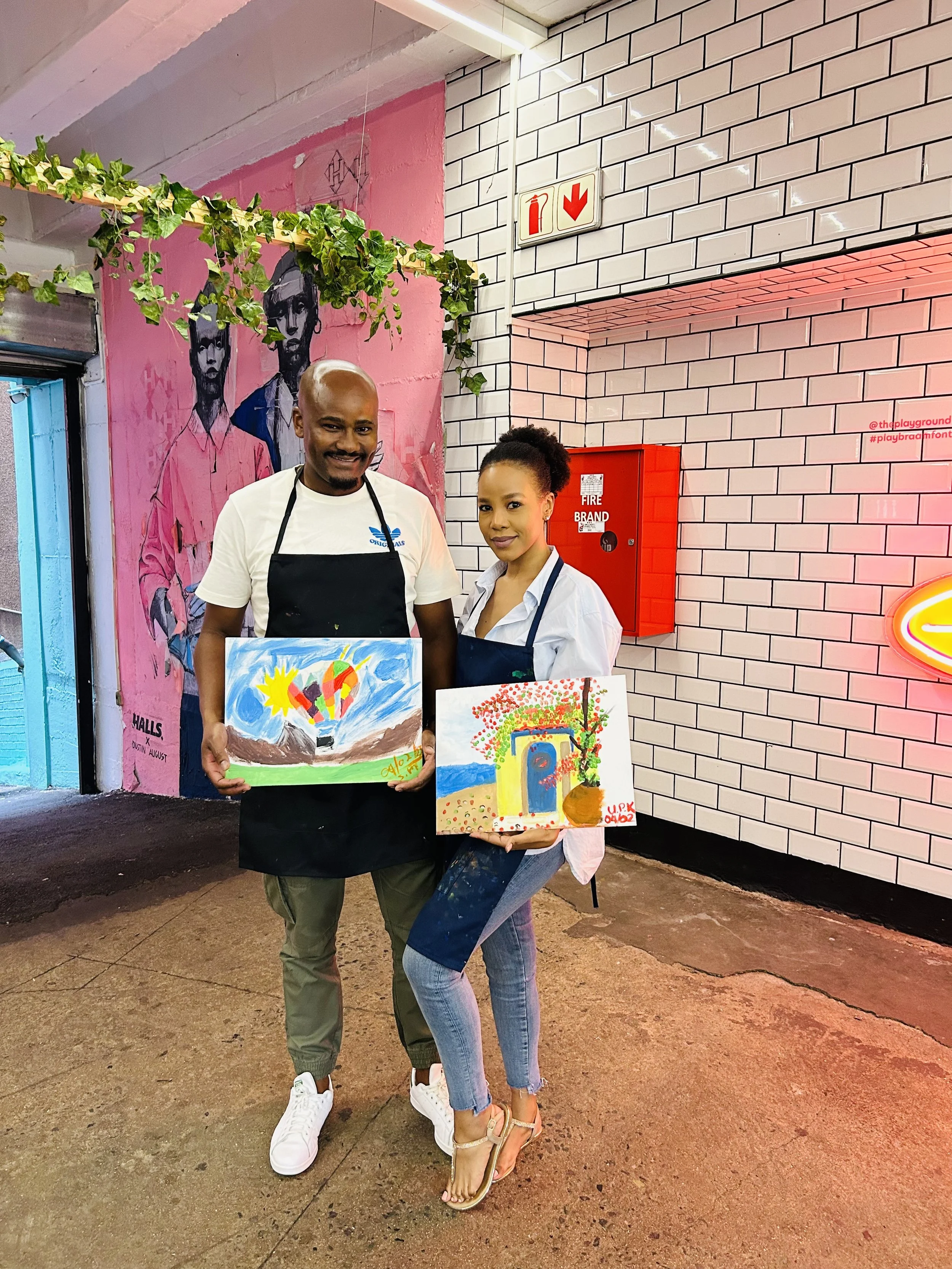 A man and woman standing side by side in an urban setting holding colorful paintings. The man is wearing a white t-shirt, a black apron, and khaki pants. The woman is dressed in a white blouse, blue jeans, and beige high-heeled sandals. Behind them a