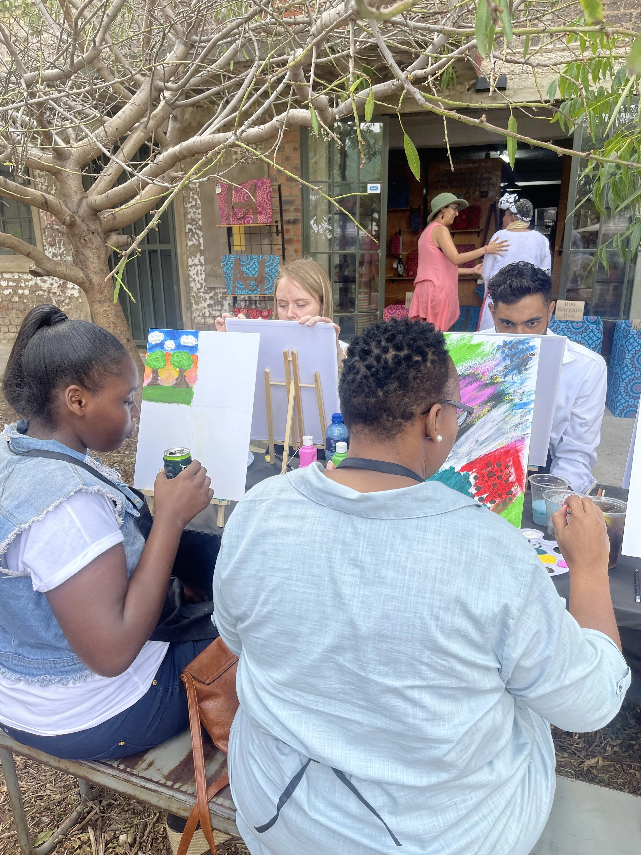 A group of people participating in an outdoor painting activity under a tree, with some holding brushes and paints, while others observe or work on their paintings.