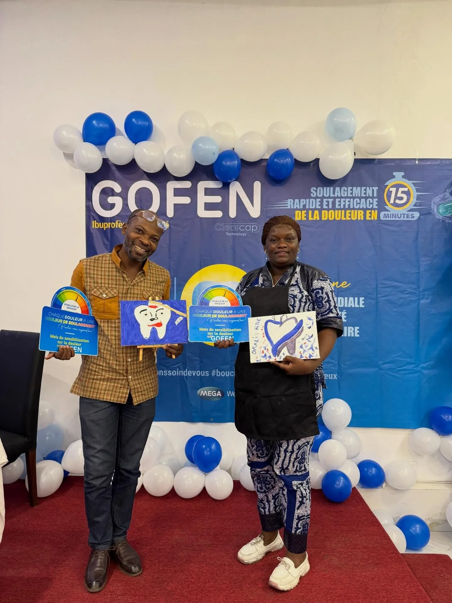 Two people standing on a red carpet at an event with blue and white balloons and a blue banner behind them. The man on the left is smiling, wearing a checked shirt and glasses on his head; the woman on the right is holding paintings, wearing a black 