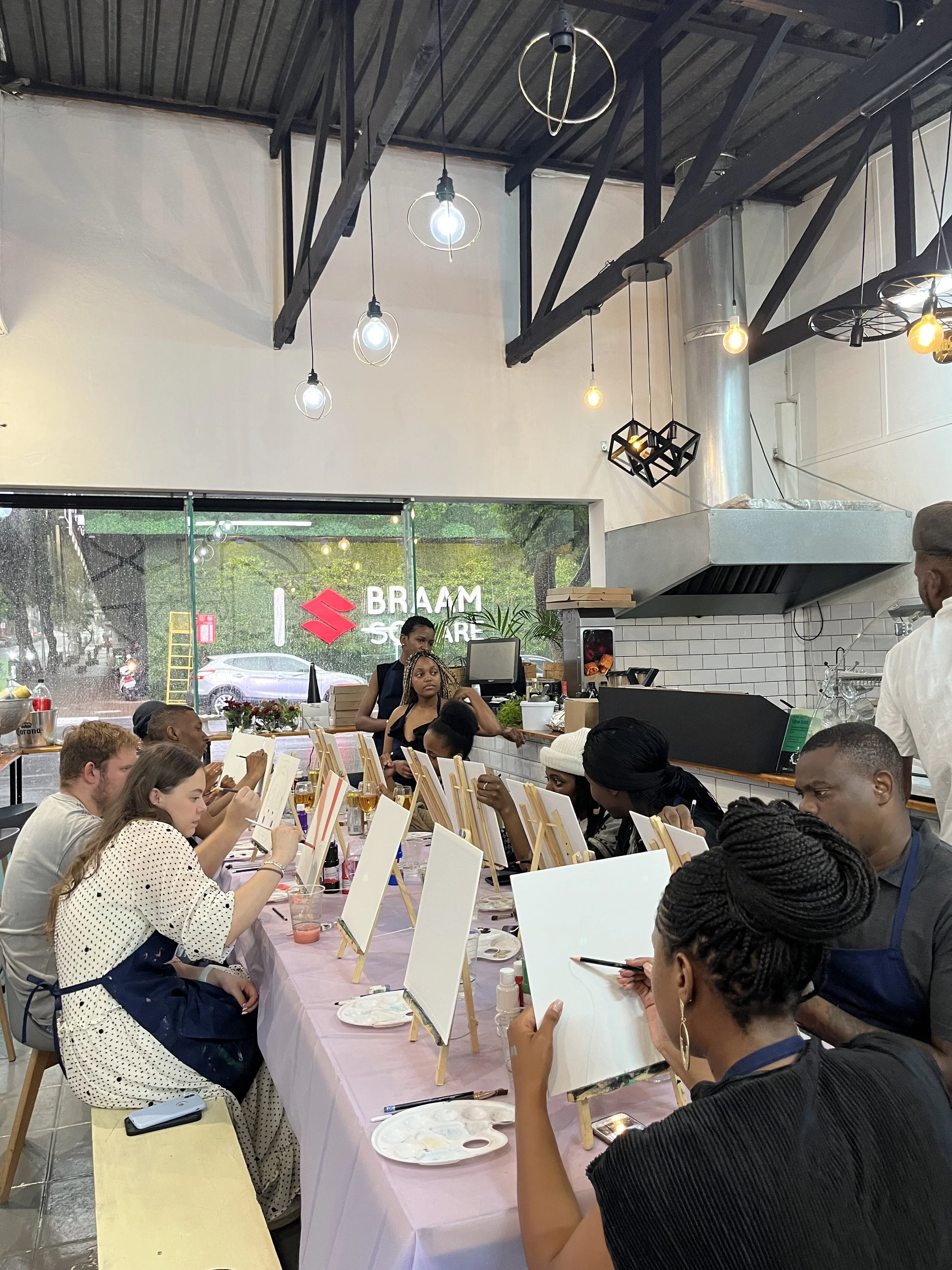 People sitting at a long table painting on canvases in a brightly lit indoor space with large windows and hanging light bulbs.