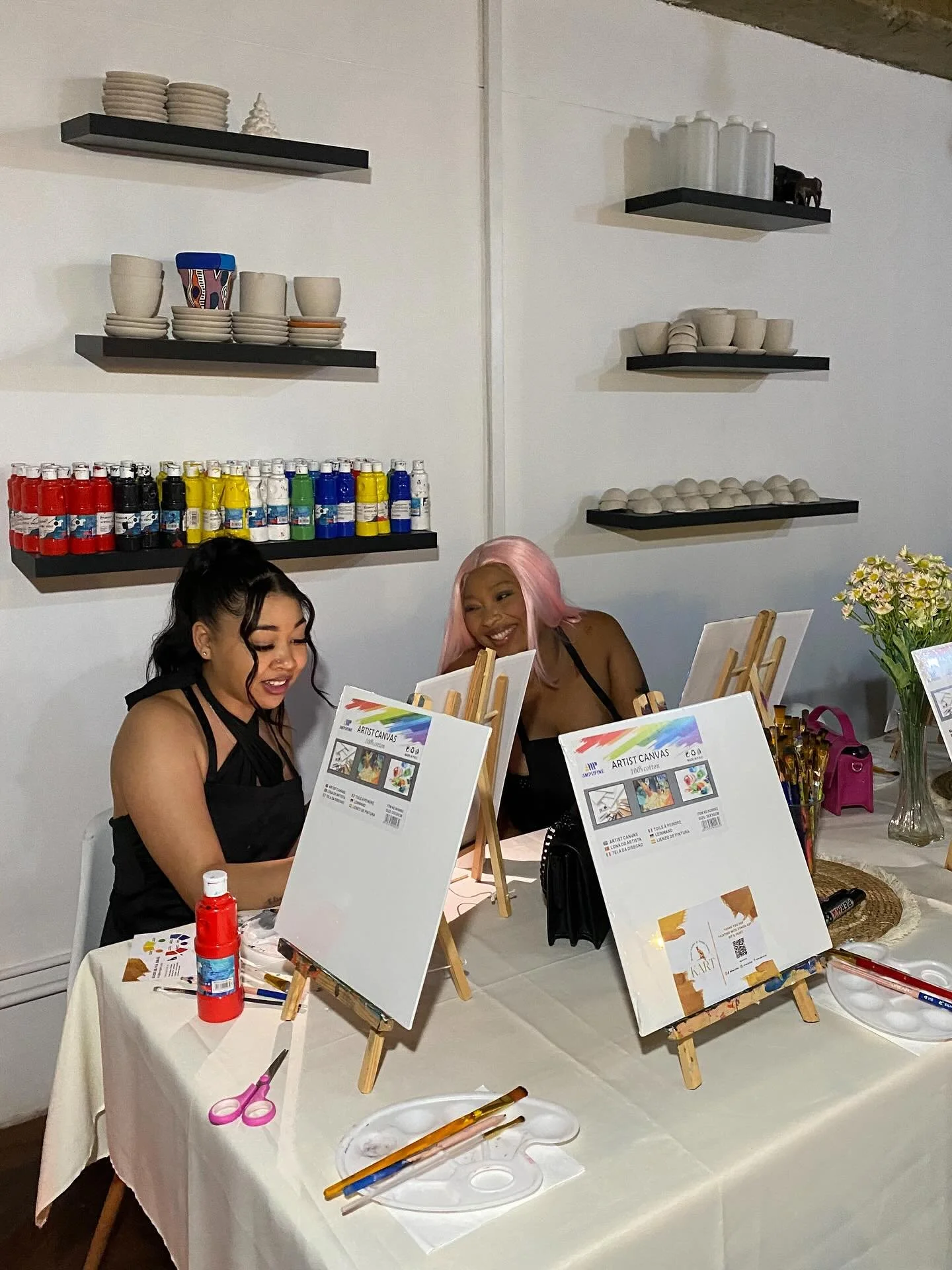 Two women sit at a table with art supplies, creating paintings on canvases supported by small easels. The table holds paint brushes, scissors, and bottles of paint.