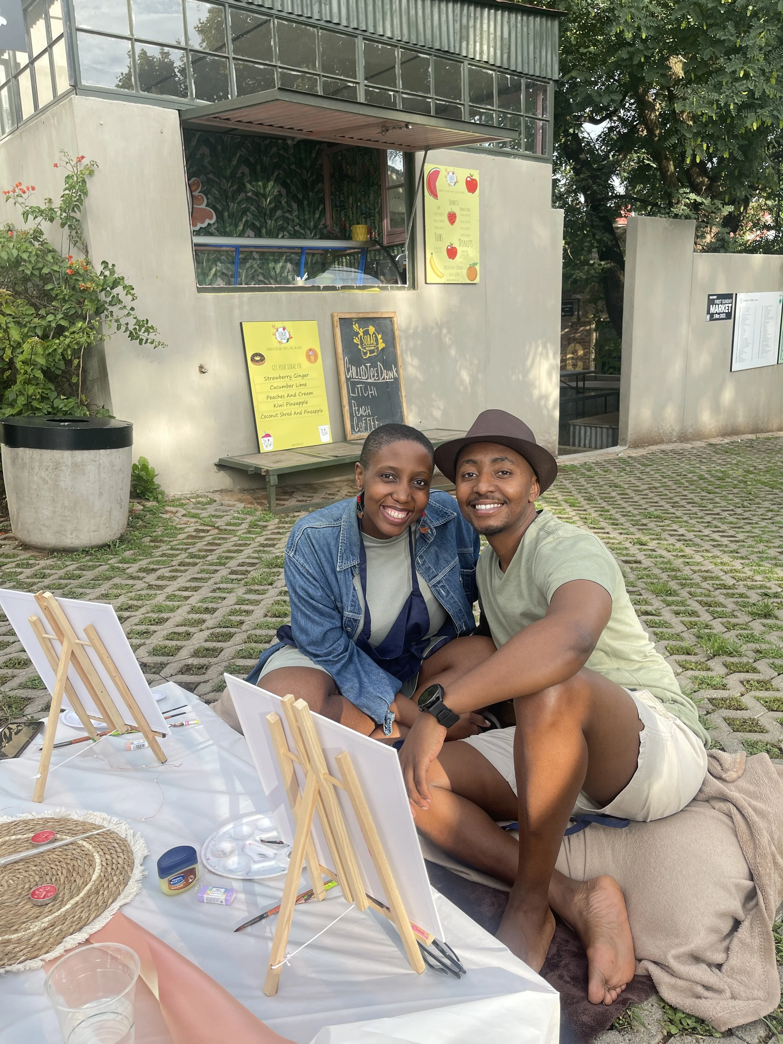 A smiling couple sitting outdoors near a table with painting supplies, with a building and trees in the background.