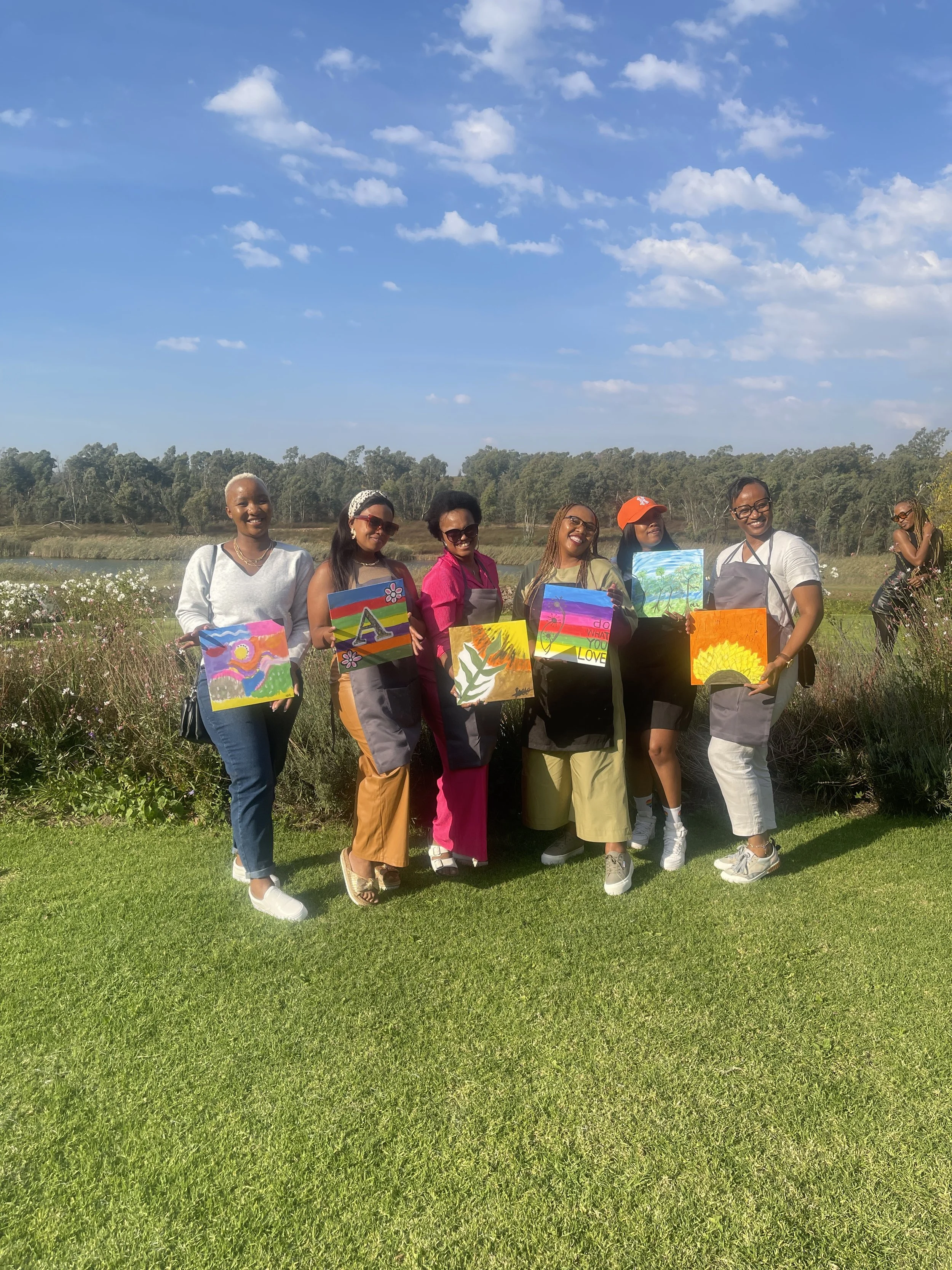 A group of seven women standing outdoors on green grass, holding colorful paintings, in front of trees and a river under a blue sky with scattered clouds.