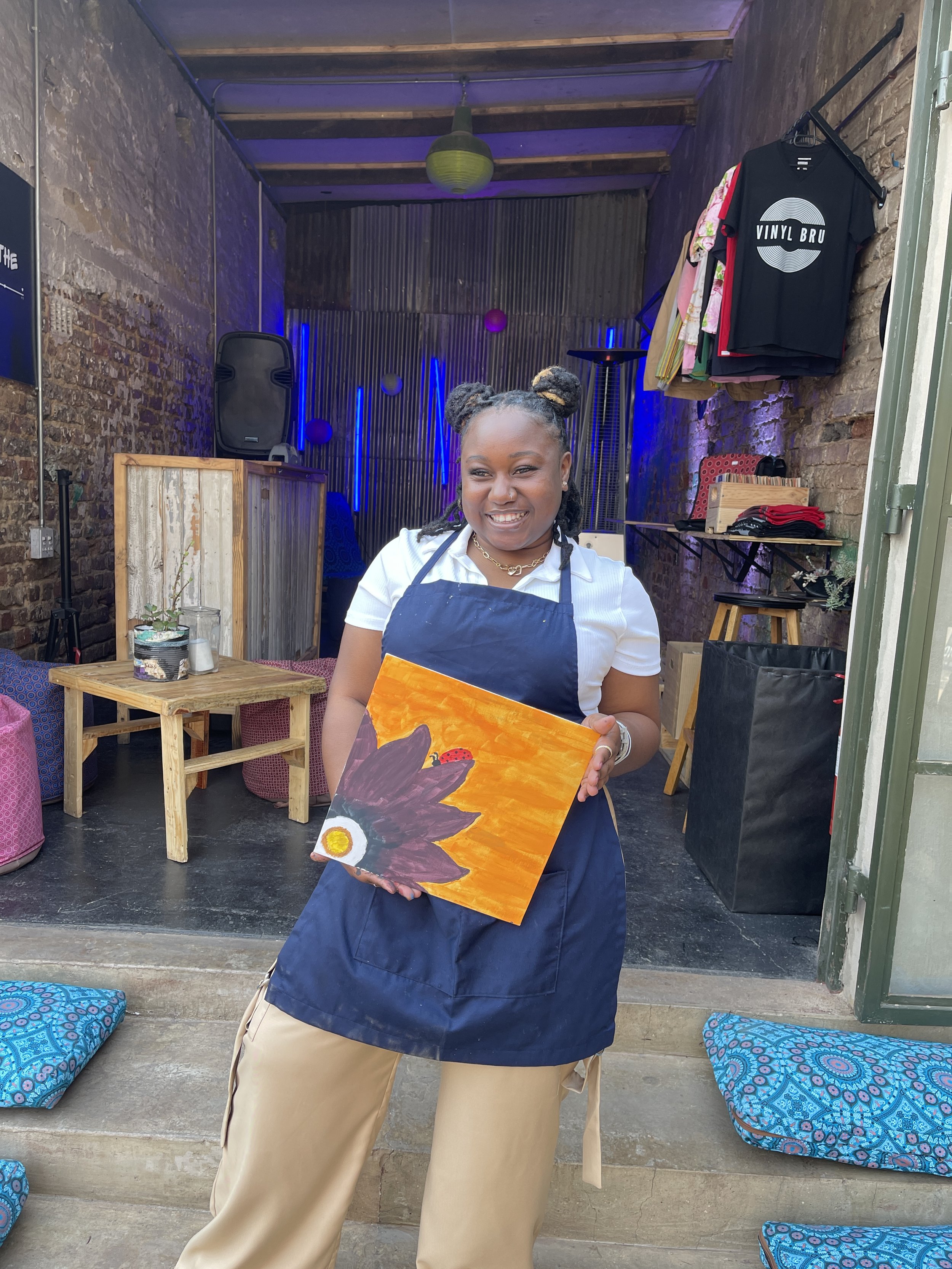 A woman standing in front of a brick wall holding a colorful painting of an eagle with a sunset background, wearing a white shirt, navy apron, and beige pants, with her hair in two buns.