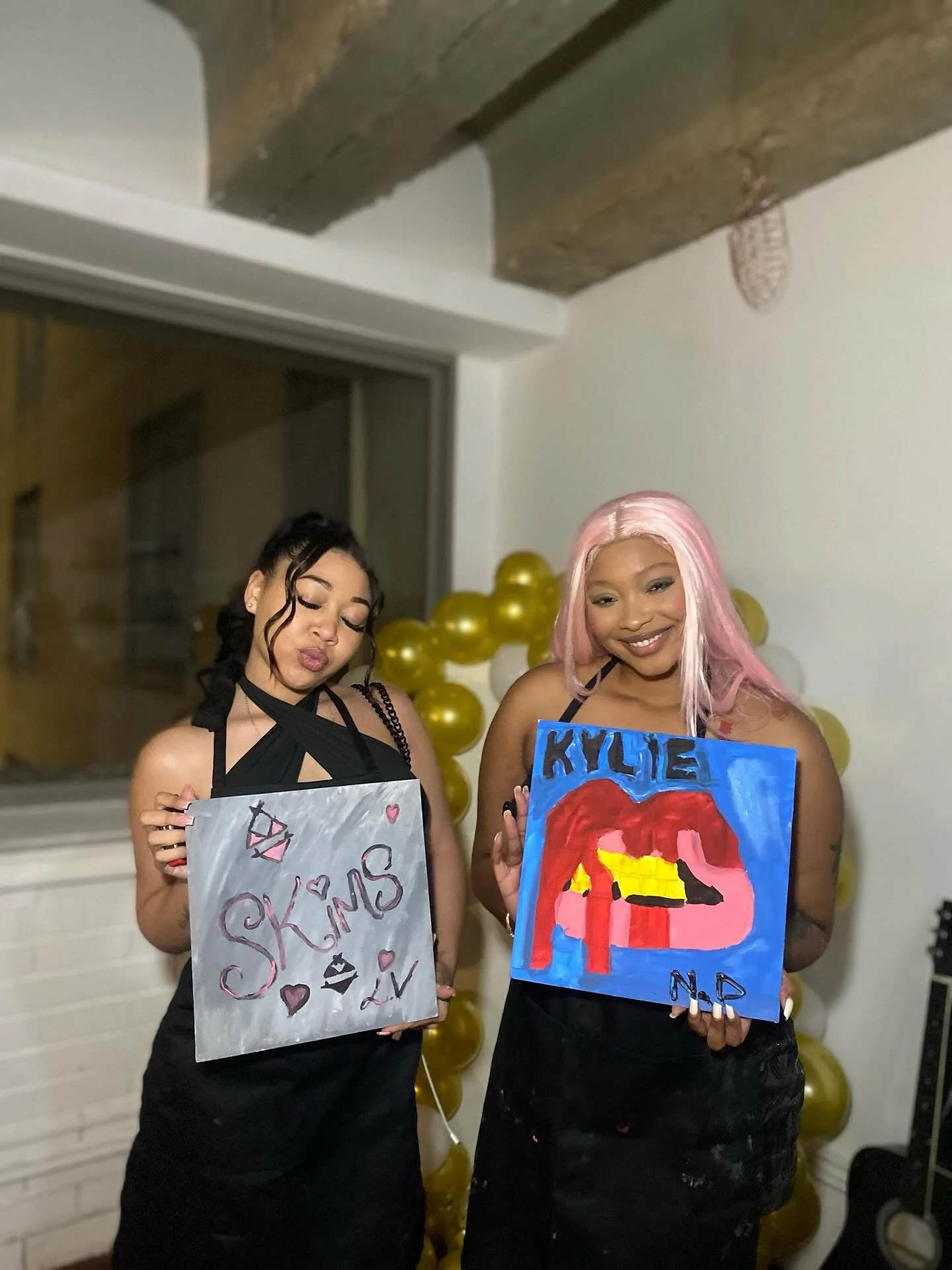 Two women standing in front of yellow balloons holding painted signs. The woman on the left has dark hair and is making a kissy face, holding a white sign with pink and black writing that says 'Kylie Skies' with hearts and lips. The woman on the righ