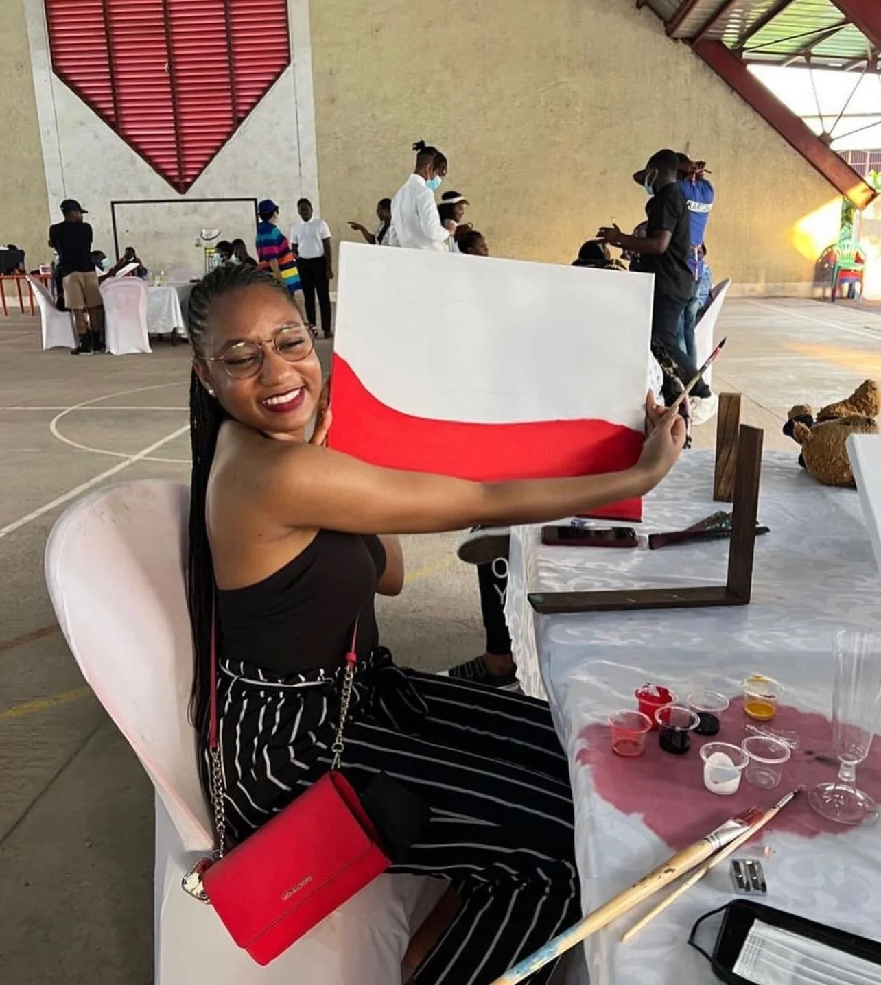 A woman with glasses and braided hair, smiling and painting an Indonesian flag on a large canvas at an indoor event. She is seated at a table with art supplies, including paints and paintbrushes, in a spacious hall with other people in the background.