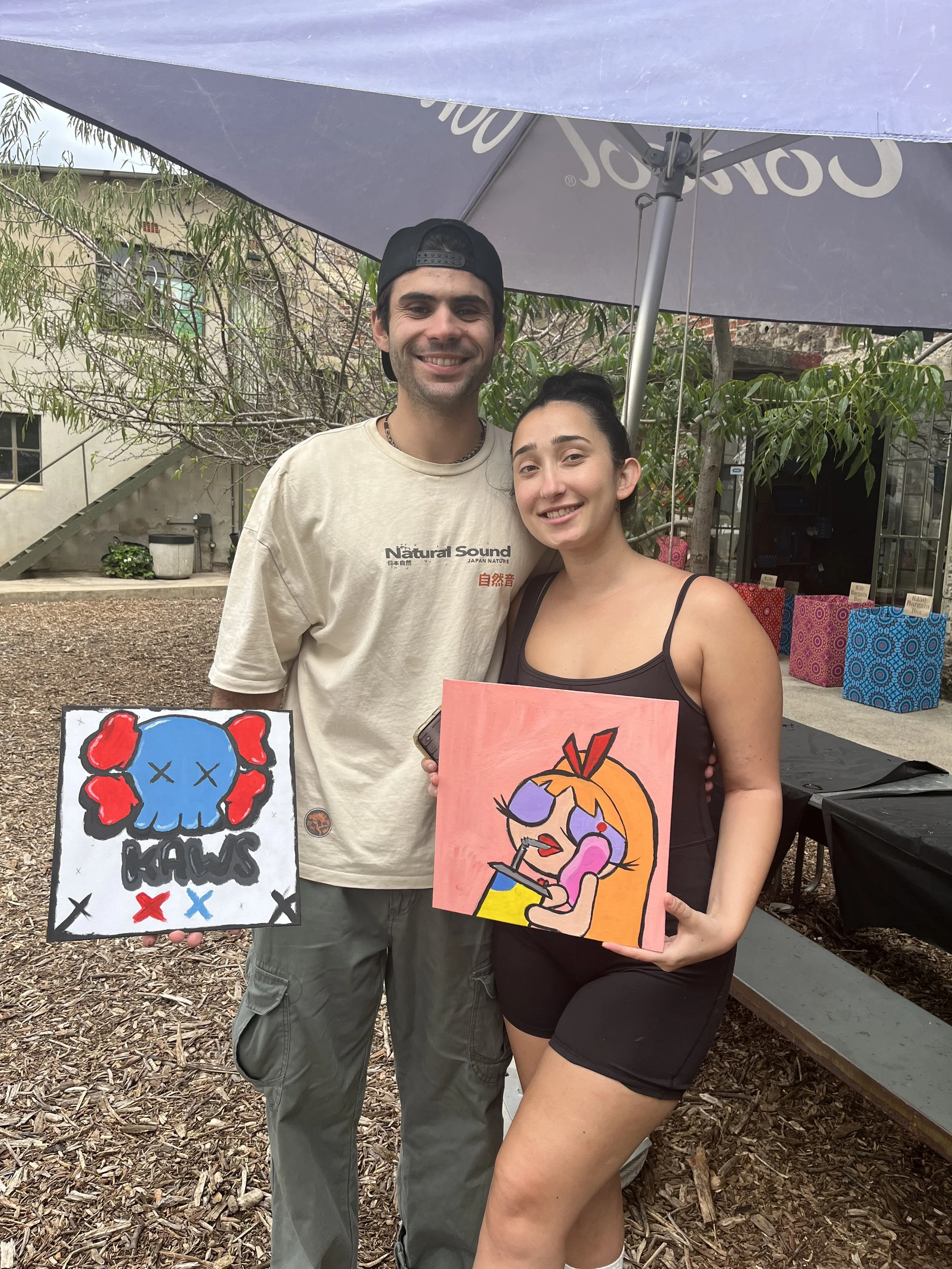 A man and a woman standing under a large purple umbrella outdoors, holding colorful artwork featuring cartoon-style characters. The man is wearing a beige T-shirt with text, a backwards black cap, and cargo pants, and is smiling at the camera. The wo
