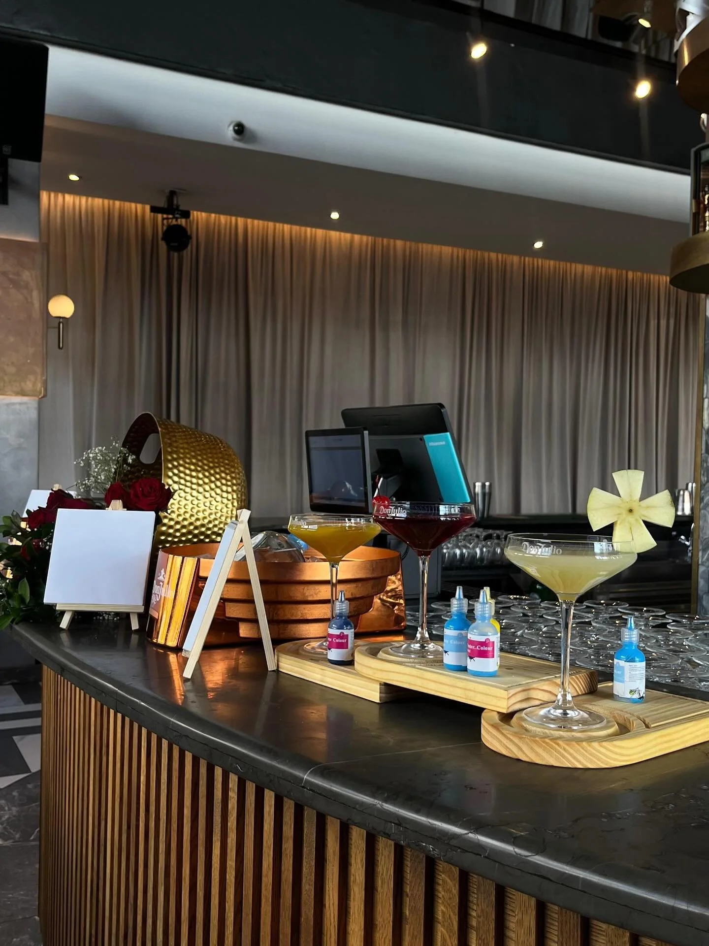 Bar counter with colorful cocktail drinks, small bottles of liquid color, a golden textured decorative object, roses, and a blank sign, with beige curtains in the background.