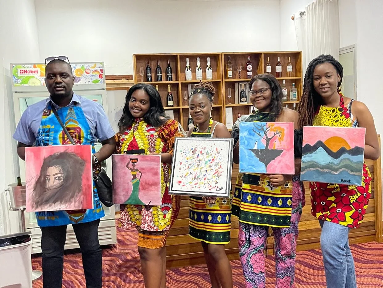 Five people, four women and one man, standing indoors in front of a wall with wooden shelves filled with wine bottles. They are holding colorful paintings and smiling. The floor is patterned with red and black stripes.