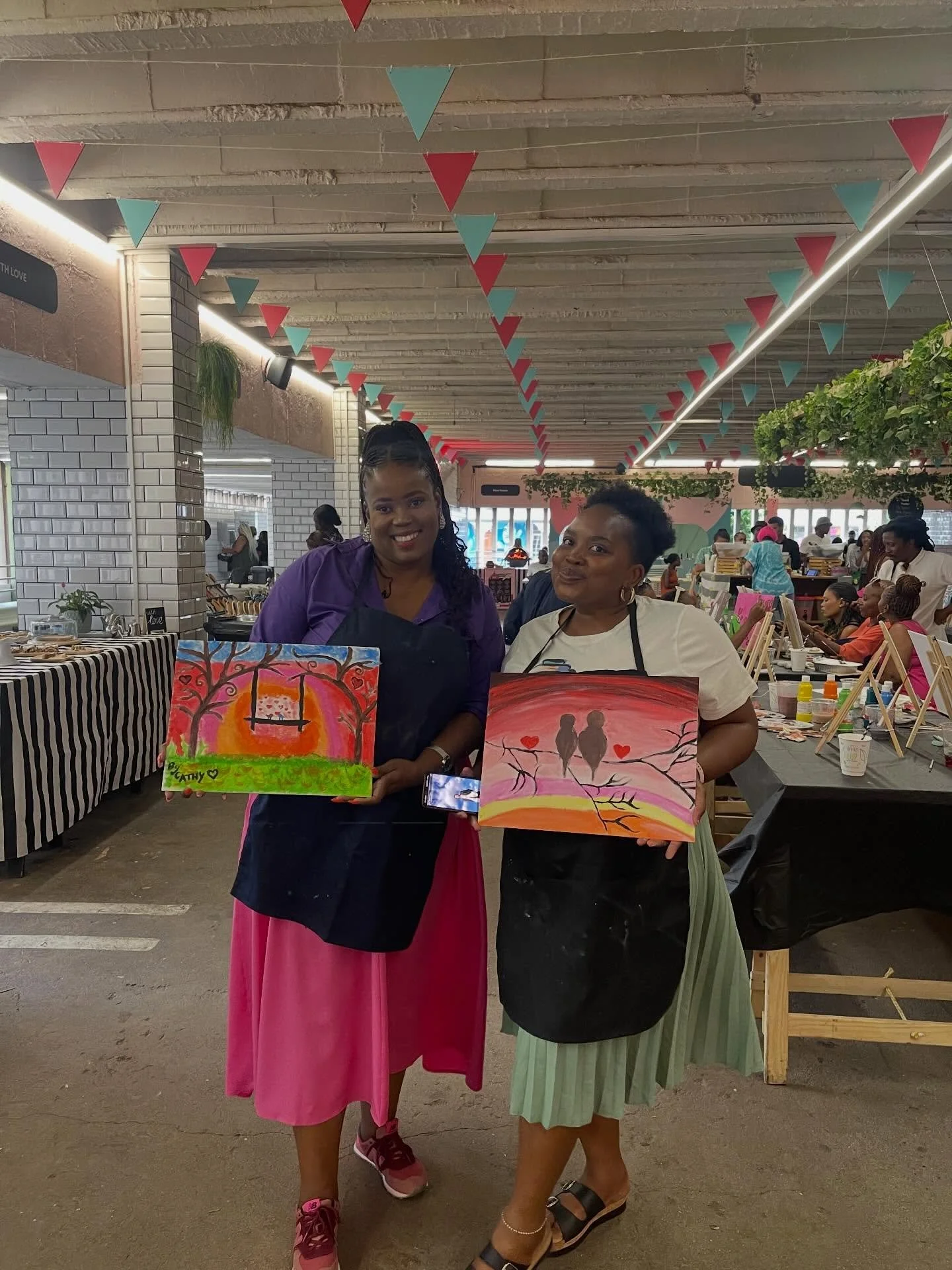 Two women stand together in a busy indoor market or craft fair, each holding colorful paintings. The woman on the left wears a purple top and long pink skirt, while the woman on the right wears a white shirt and green striped skirt. There are tables 