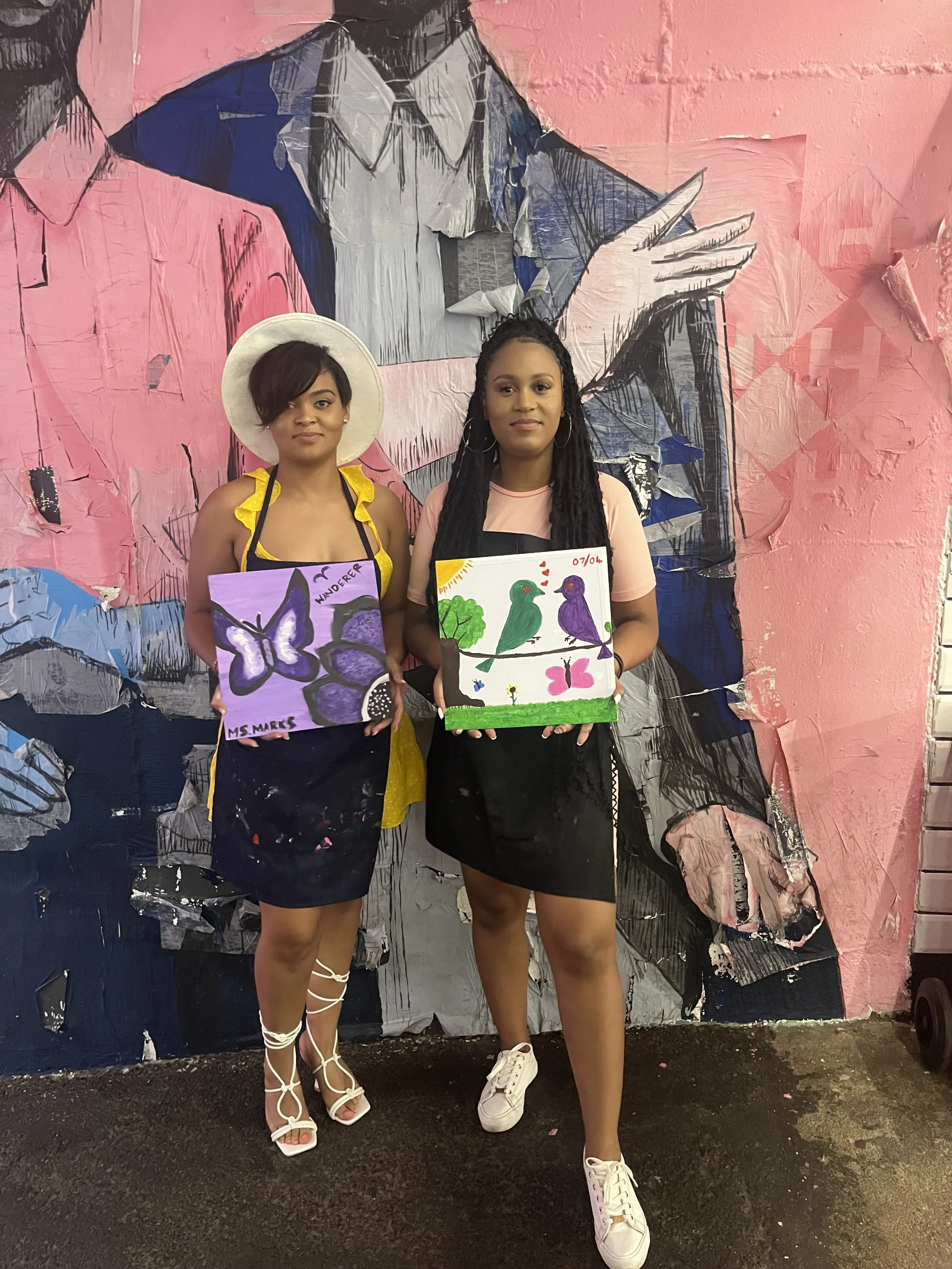 Two women standing in front of a colorful, textured mural. Each woman is holding a painting of nature scenes with birds and butterflies.