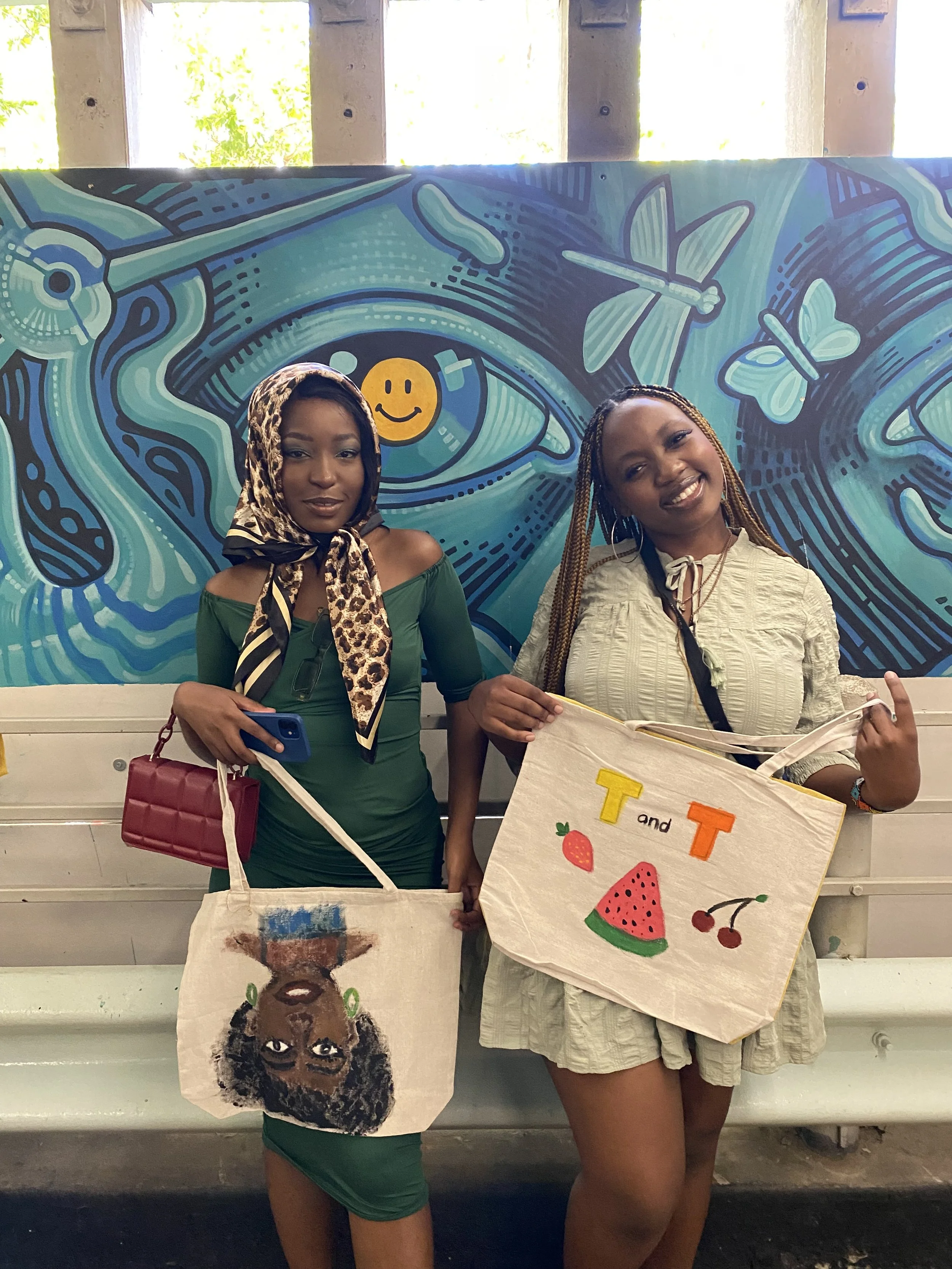 Two women stand outdoors with a colorful mural behind them. Each holds a painted tote bag with artistic designs and illustrations.
