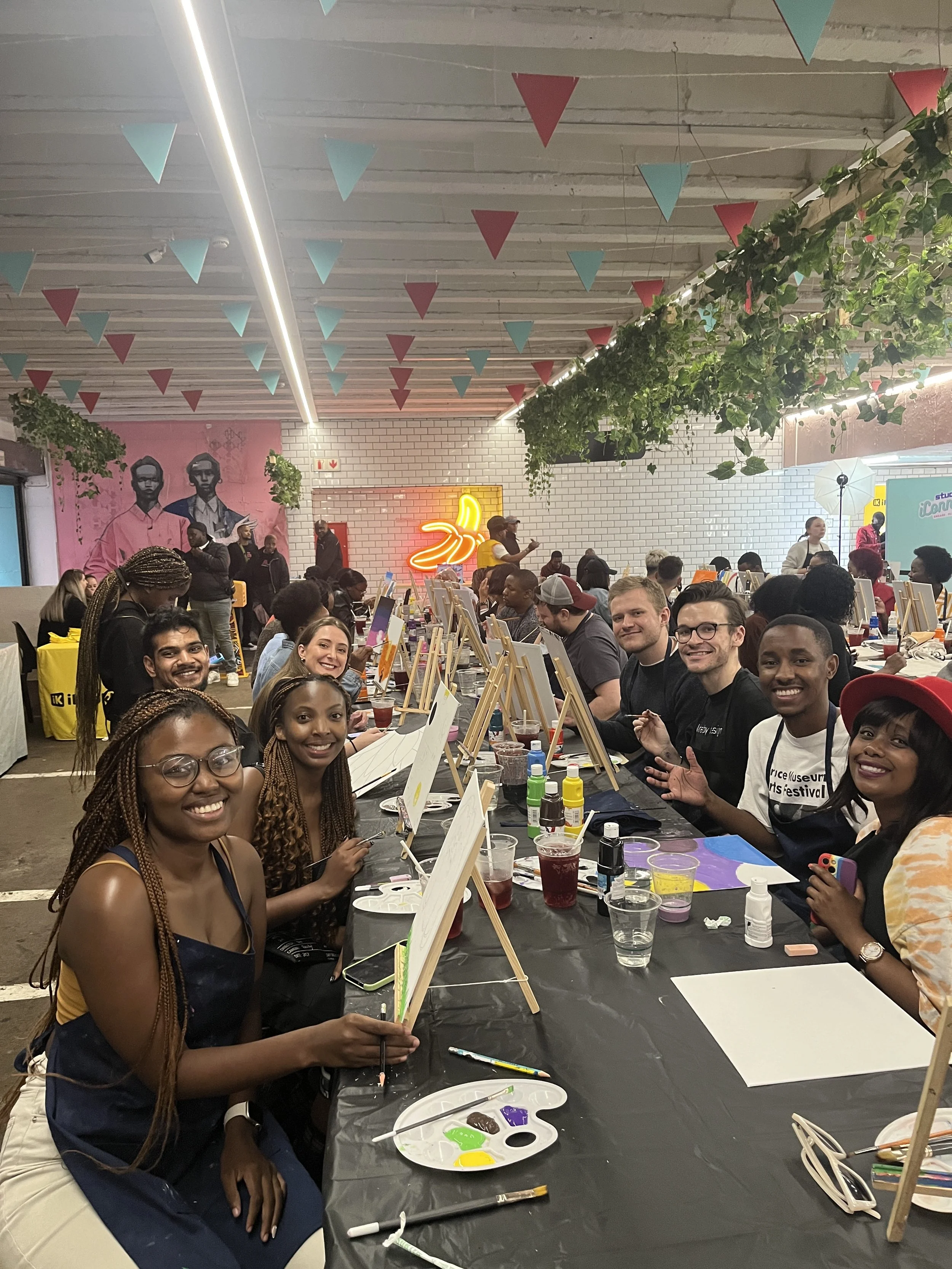 People seated at long table with painting supplies, including paint and brushes, engaging in a group art activity in a decorated indoor space with hanging flags and wall art.